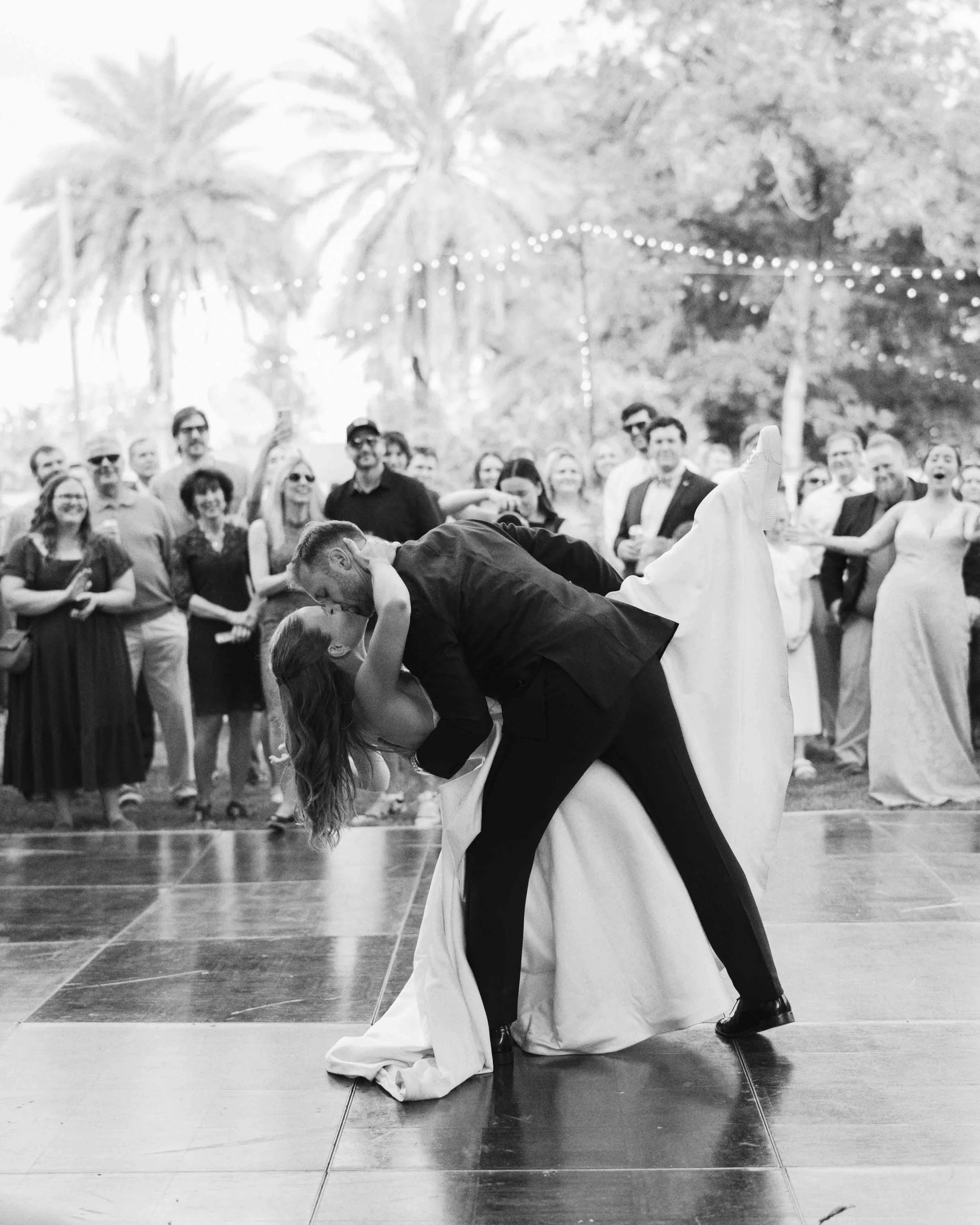 A couple dancing closely on a dance floor while a crowd of people watch and take pictures in the background, outdoors with string lights and palm trees.