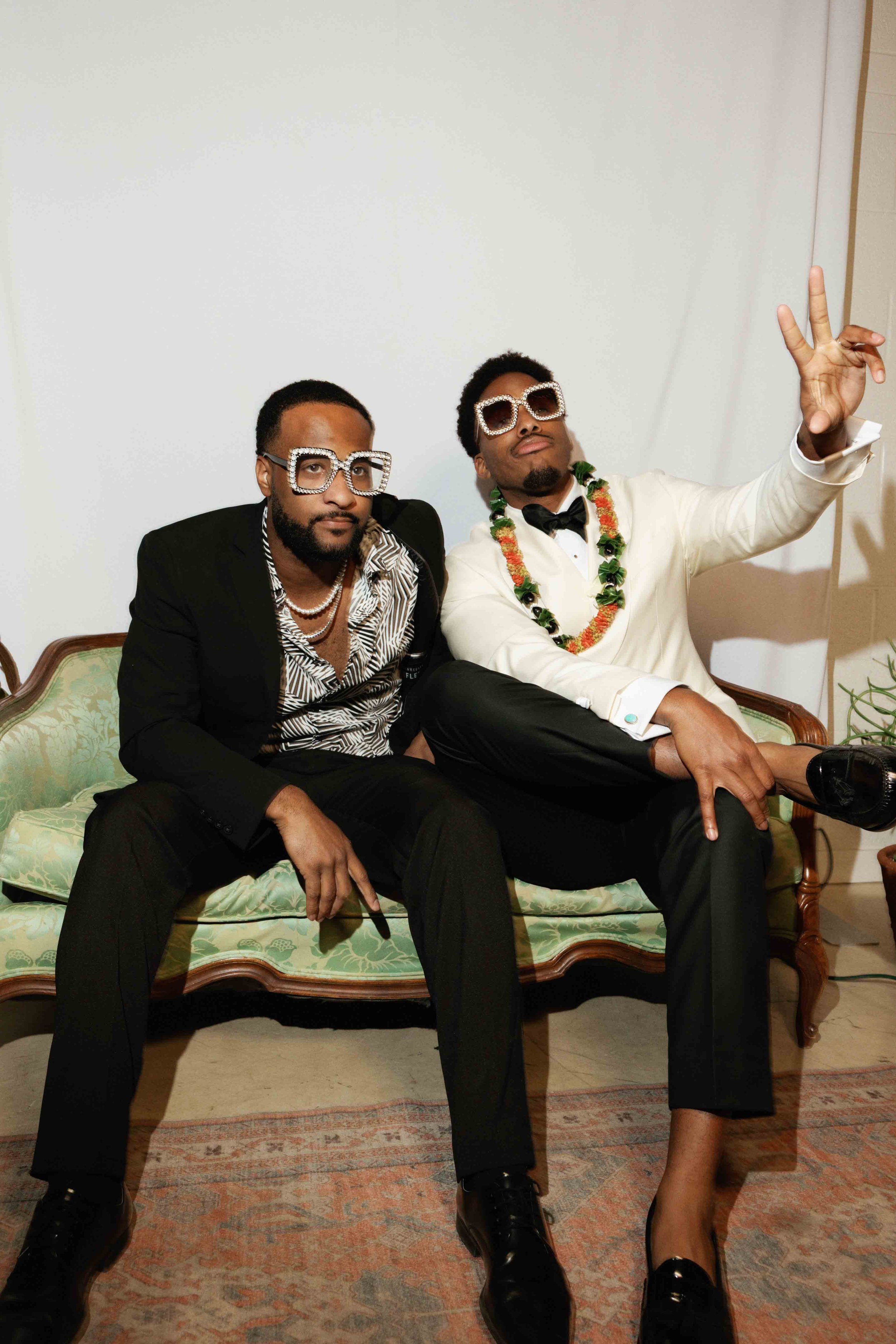 Two men in formal attire sitting on a vintage sofa, wearing large sunglasses and jewelry, with their legs crossed, one making a peace sign gesture, in a room with a white wall and patterned carpet.