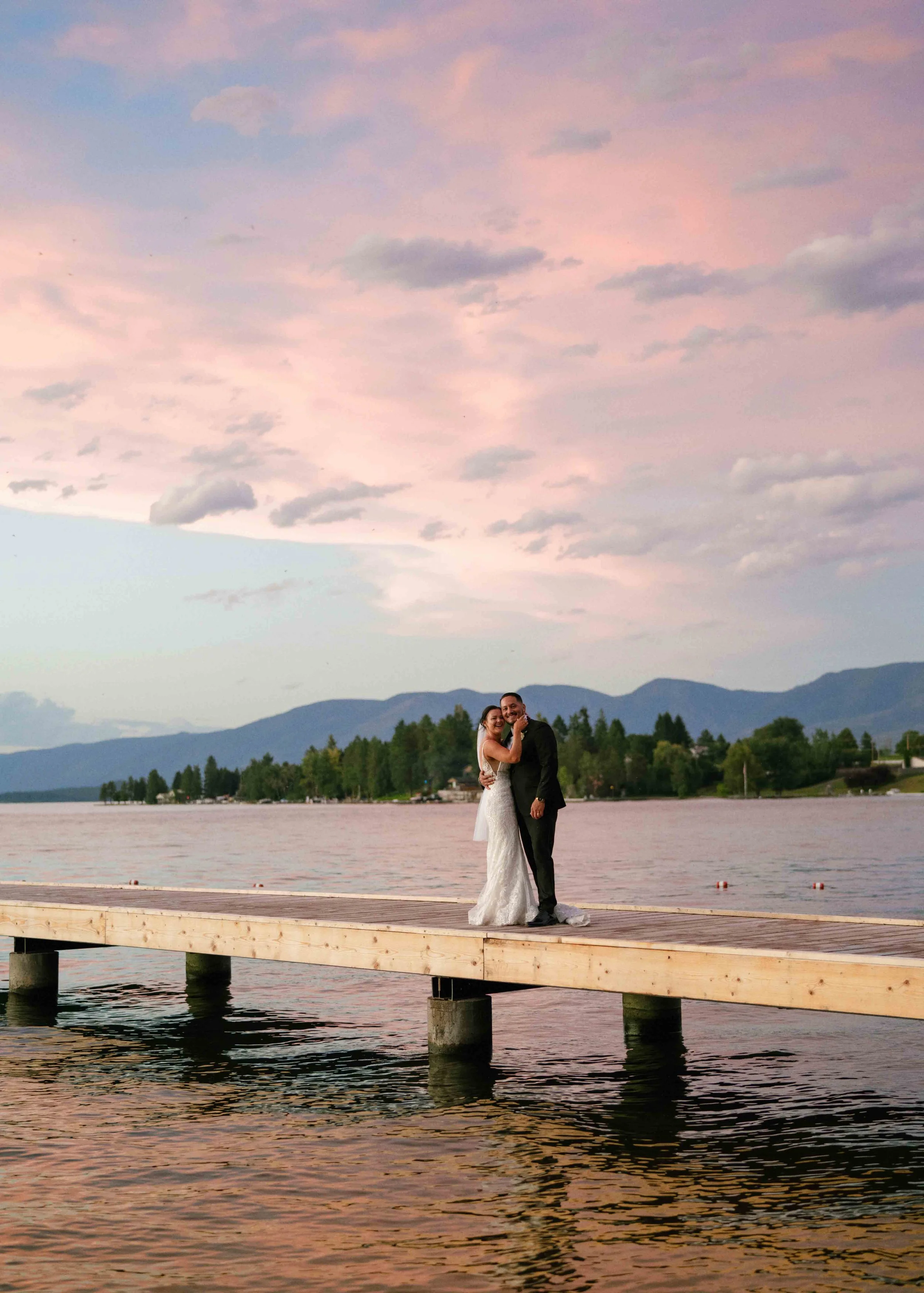 A bride and groom standing together on a wooden dock by a lake during sunset, with mountains in the background and pink clouds in the sky.