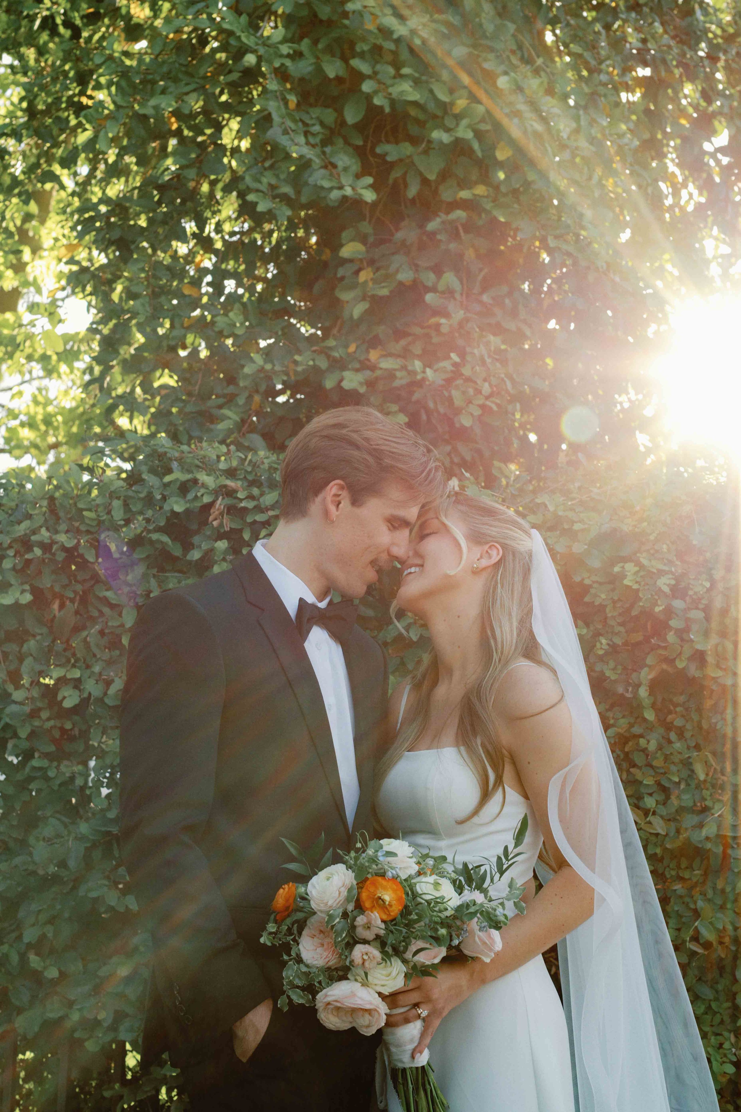 A couple in wedding attire sharing a close moment outdoors, with greenery and sunlight in the background. The woman is holding a bouquet of flowers.