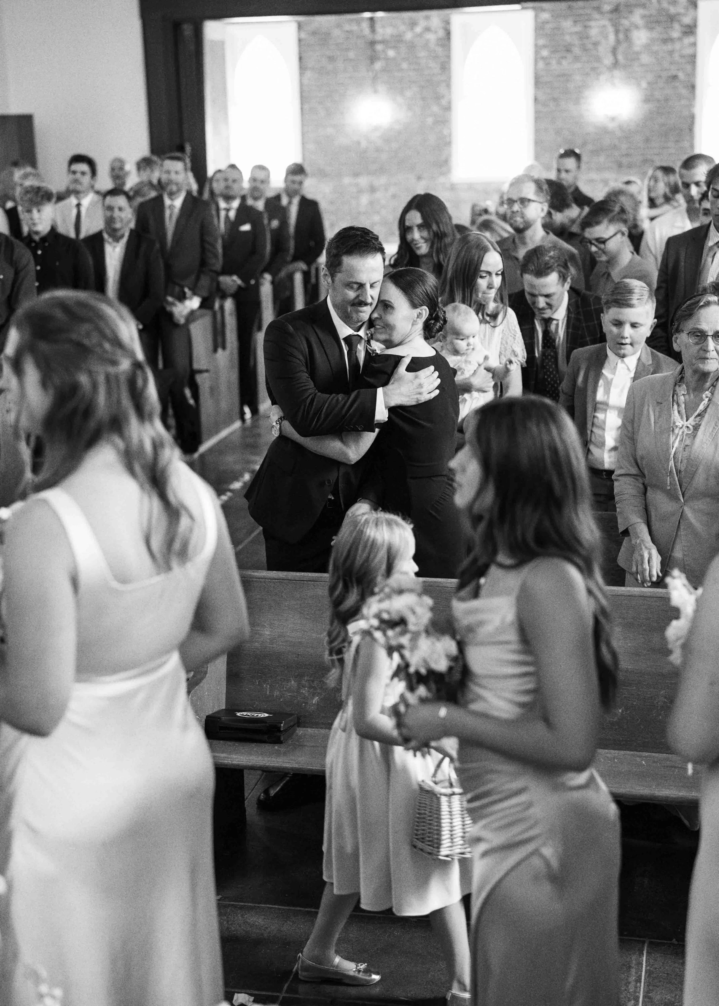 A wedding ceremony in a church with guests and children. The bride and groom are in the center, sharing a close moment while others observe. The church has brick walls and arched windows, and children are dressed in dresses and suits.