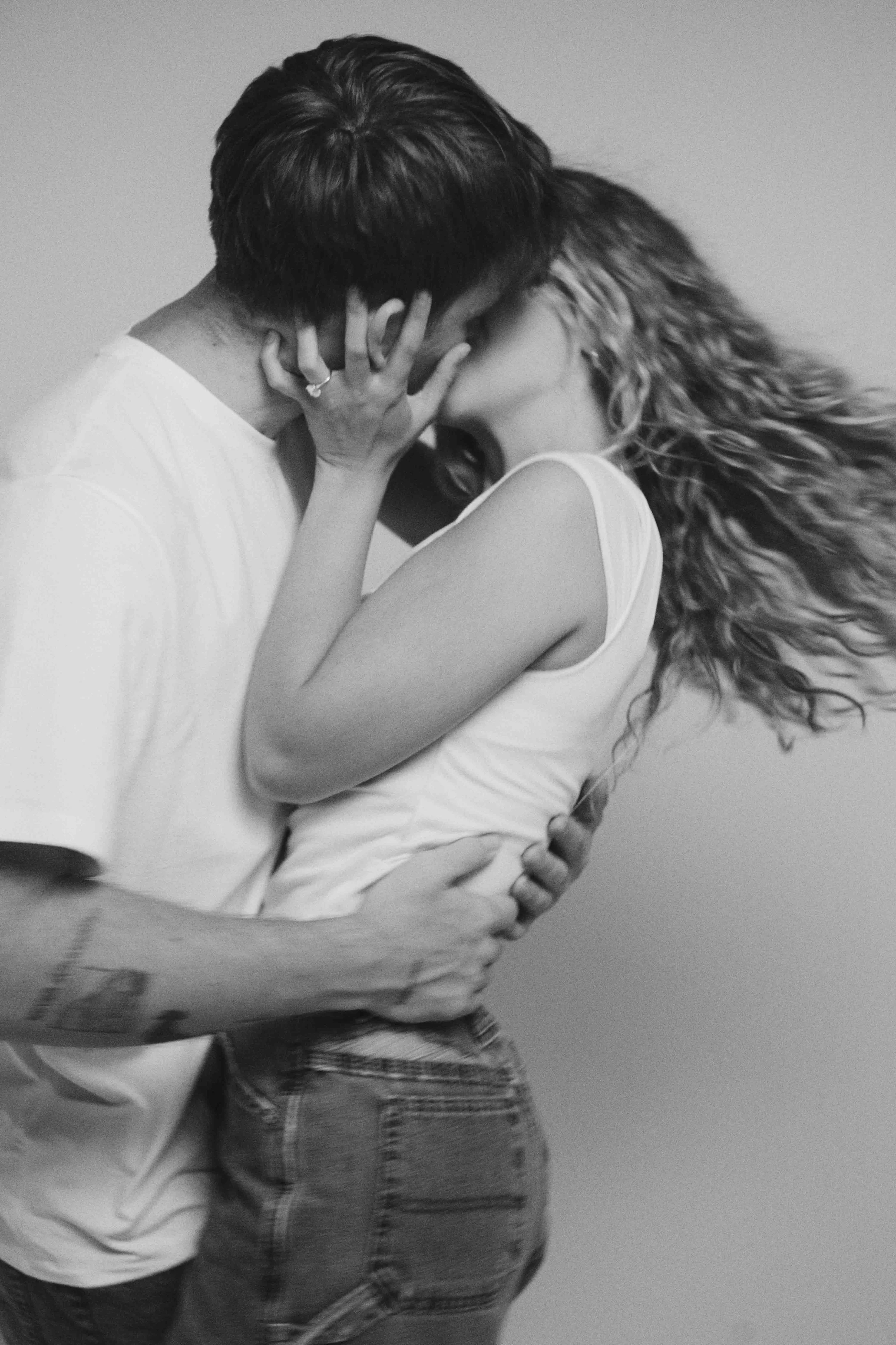 A black and white photo of a couple with their faces close together, the woman touching the man's face, and both embracing each other against a plain background.