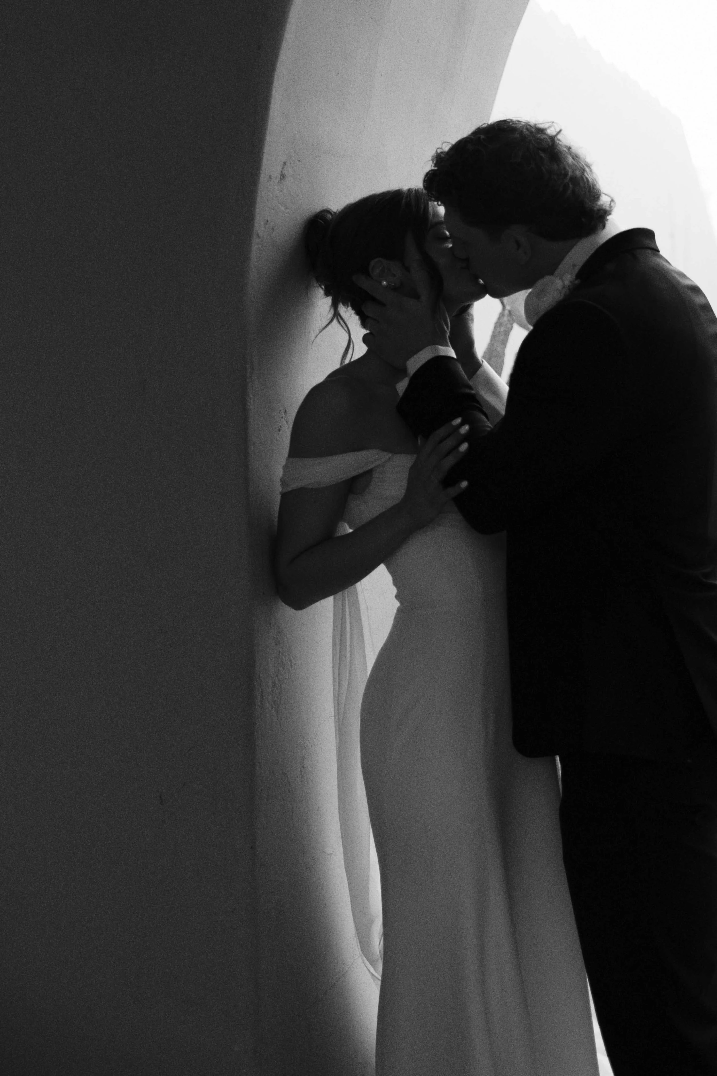 A black and white photo of a couple sharing a kiss against a wall at the beach.
