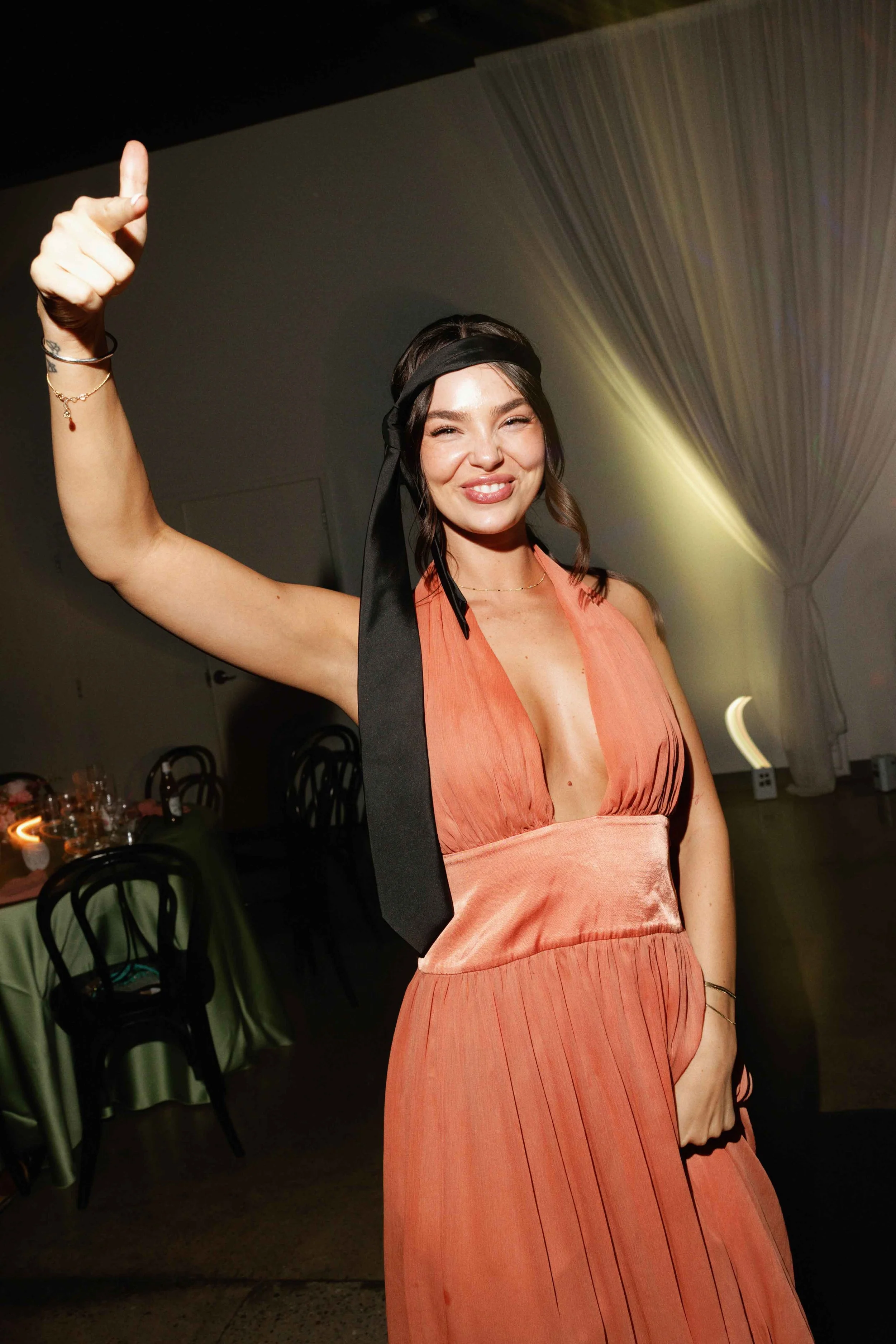 A woman wearing a coral dress and black headband smiling and giving a thumbs-up at a party or event, with tables and chairs visible in the background.