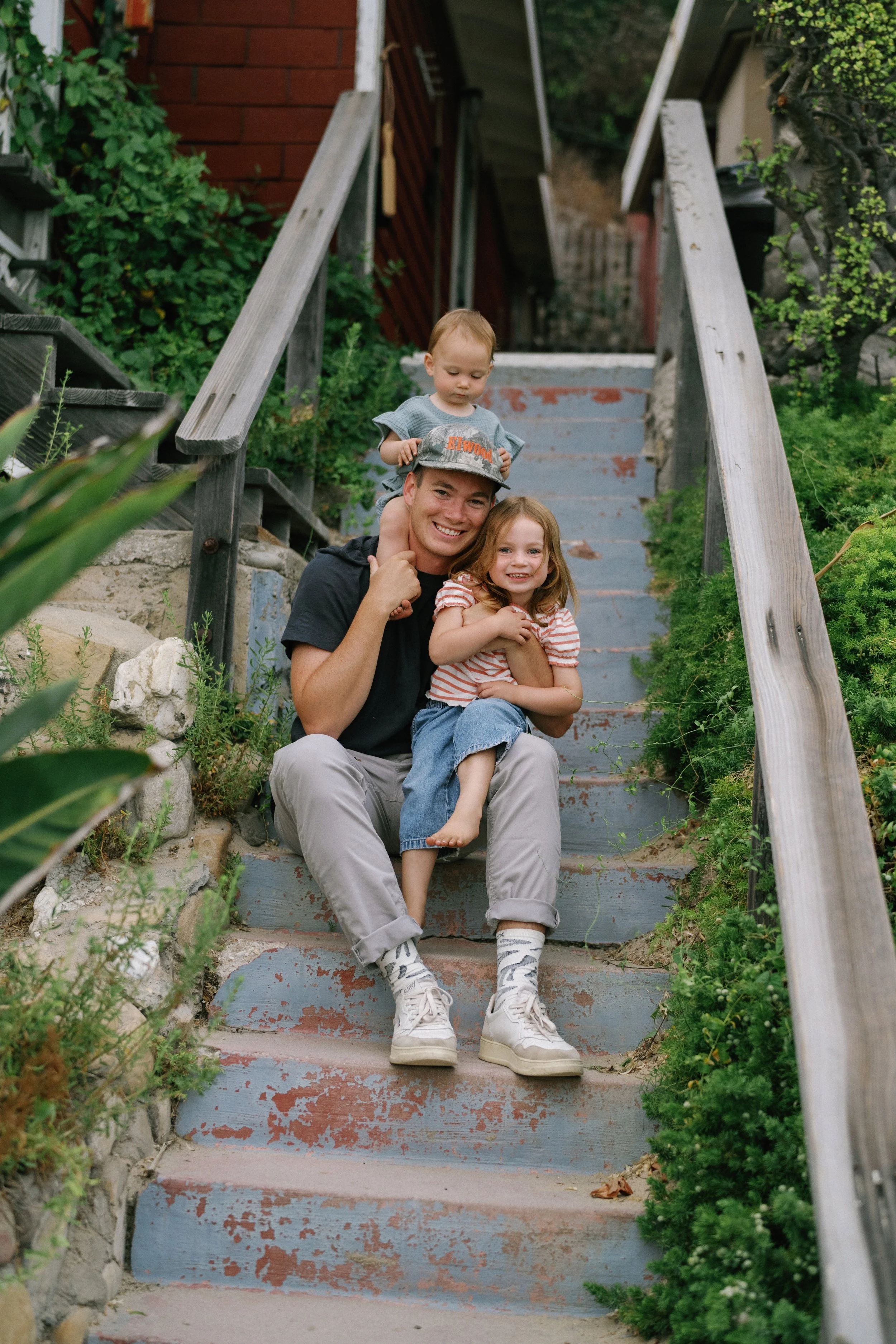 A happy man with two children sitting on outdoor stairs, surrounded by greenery.