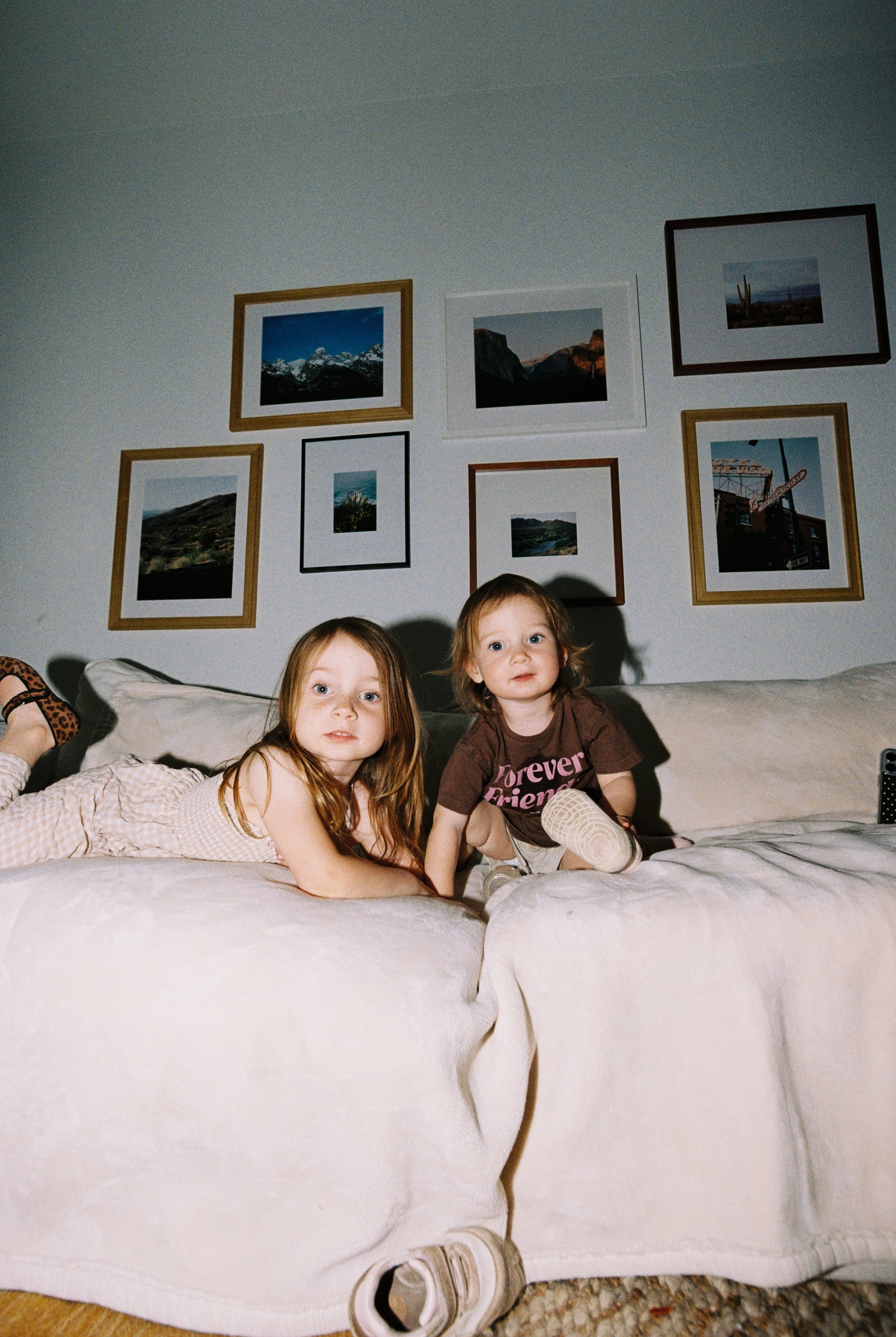 Two young girls with light skin and red hair lying on a bed with white bedding, in a room with framed landscape photographs on the wall behind them.