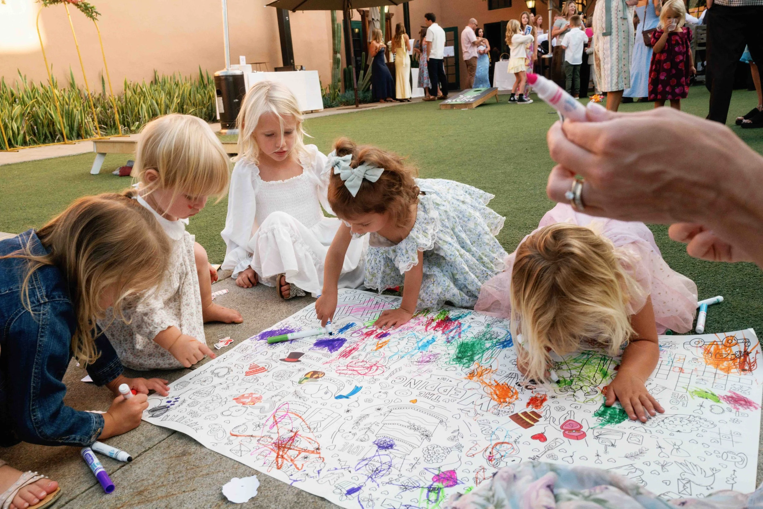 Children gather around a large, colorful poster sheet on the ground, drawing and coloring with markers at an outdoor event. In the background, there are more people mingling, a building, and some picnic tables.