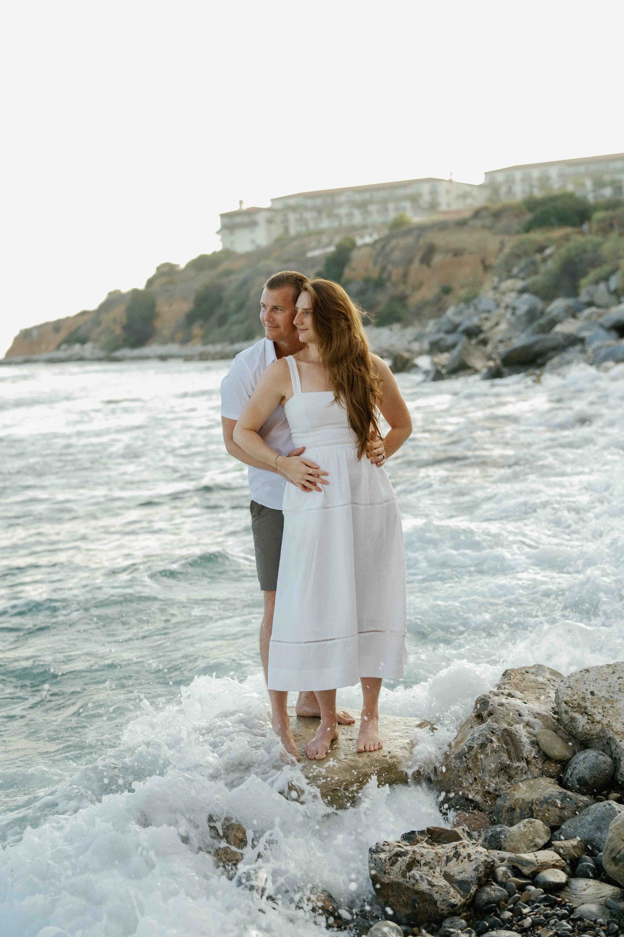 A couple standing in ocean water on rocks, with a coastal cliff and buildings in the background.
