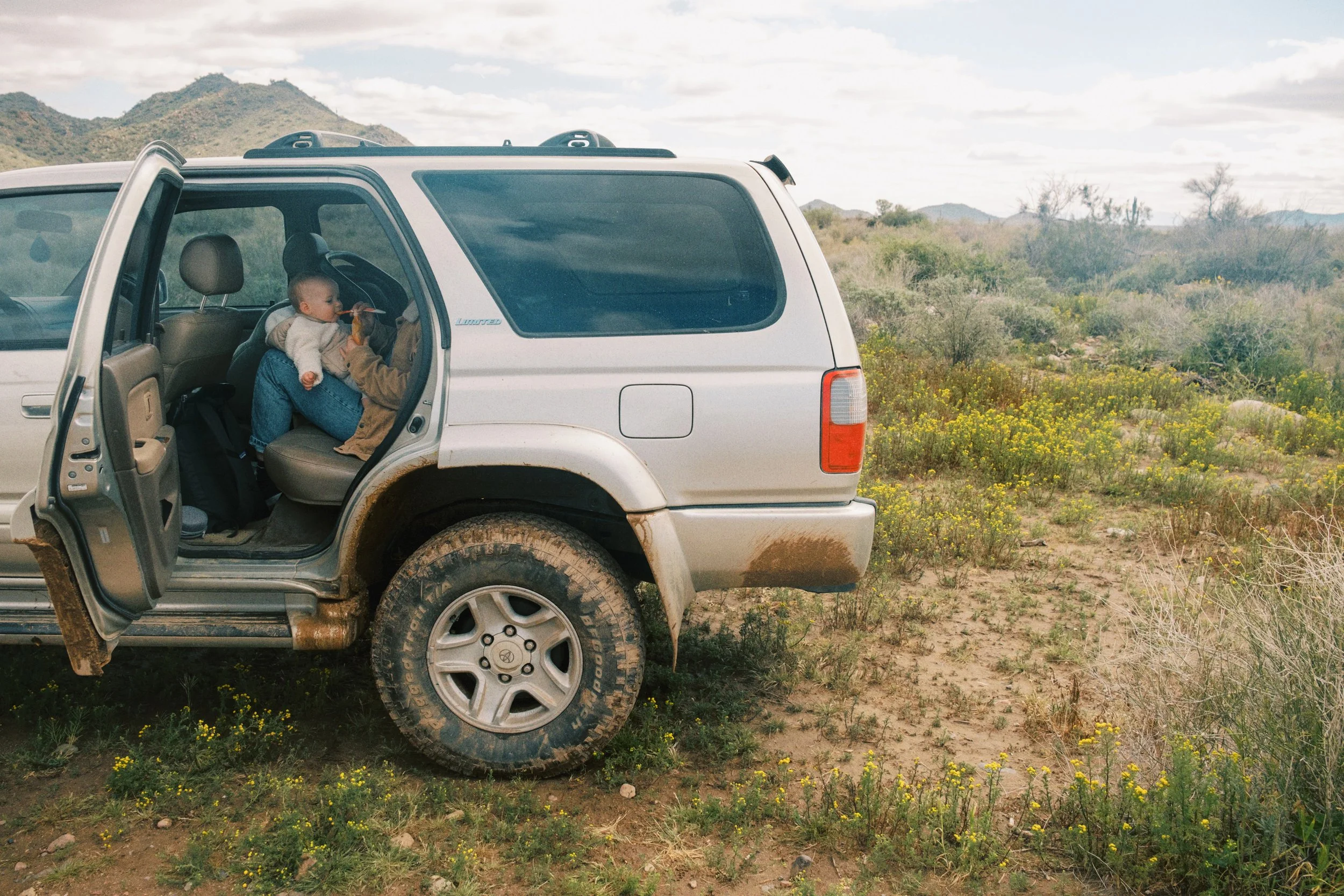 A silver SUV parked on dirt in a desert landscape with mountains and cloudy sky in the background. The rear door on the driver side is open, revealing a child in a car seat and an adult filling a cup or bottle for the child.