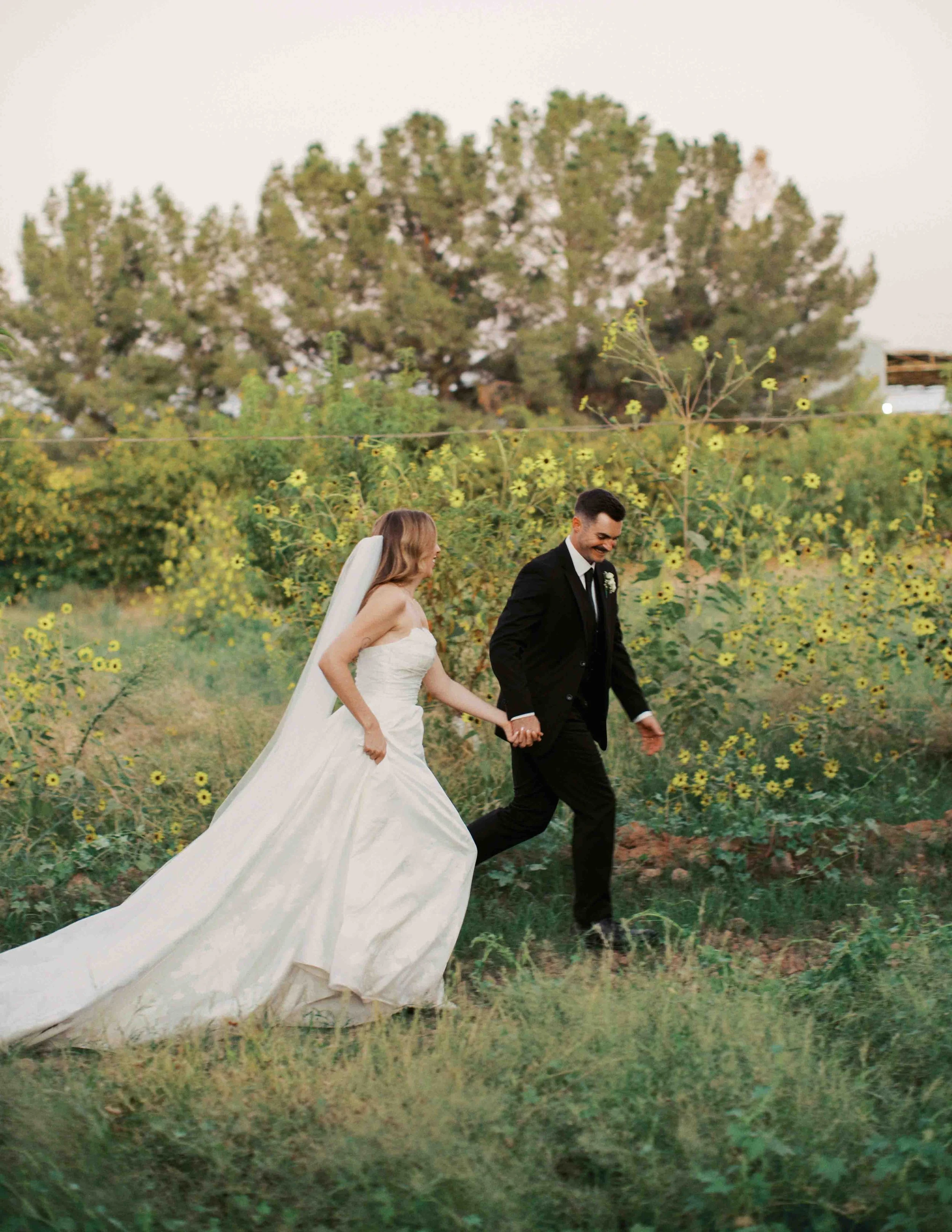 A bride and groom holding hands and walking through a field of yellow wildflowers, with a large tree and a white sky in the background, during a wedding ceremony.