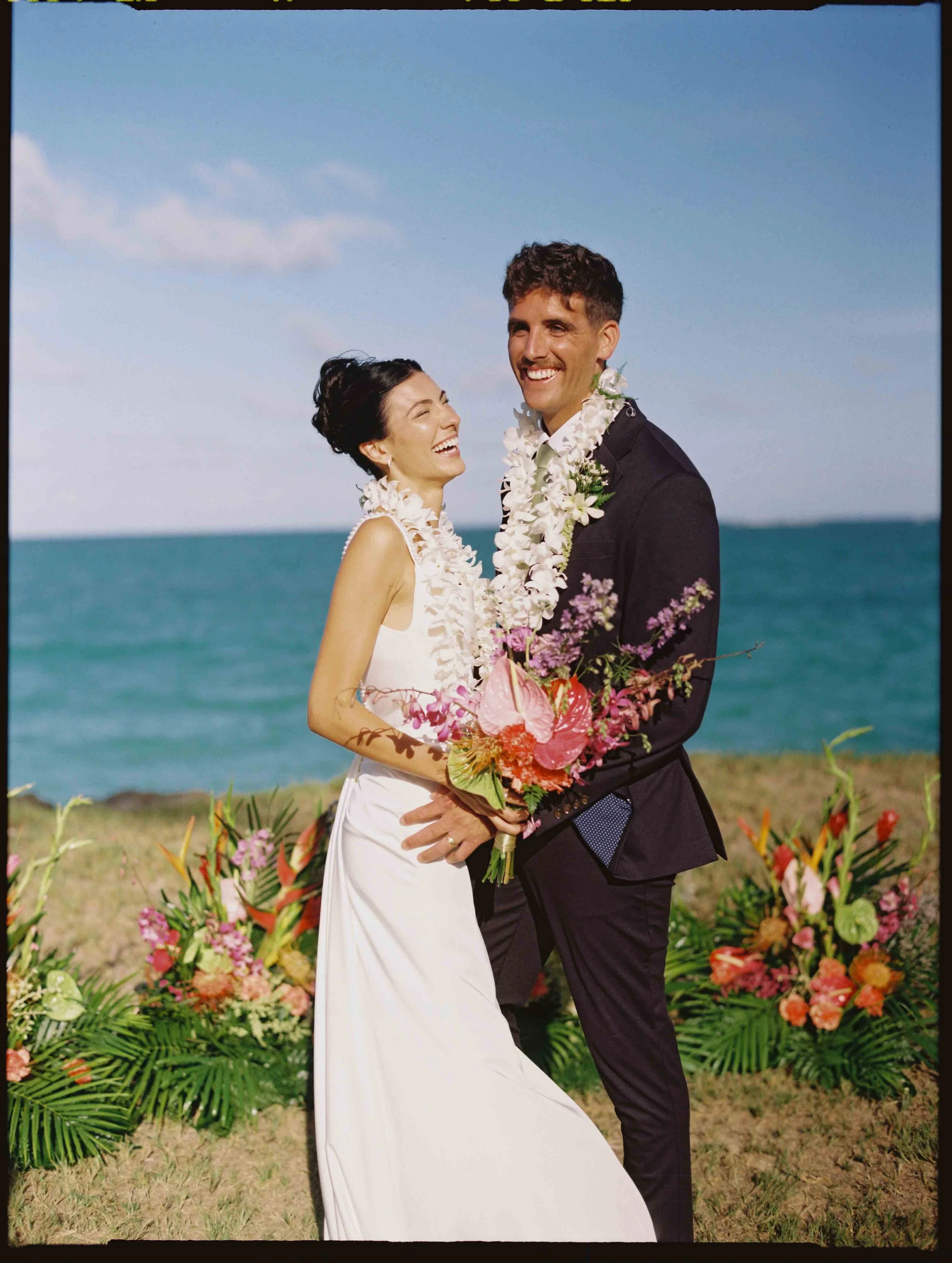 A smiling couple standing on a beach with colorful flowers and palm leaves in the foreground. The woman wears a white dress and the man wears a dark suit, both adorned with leis. The background features the ocean and a blue sky with a few clouds.