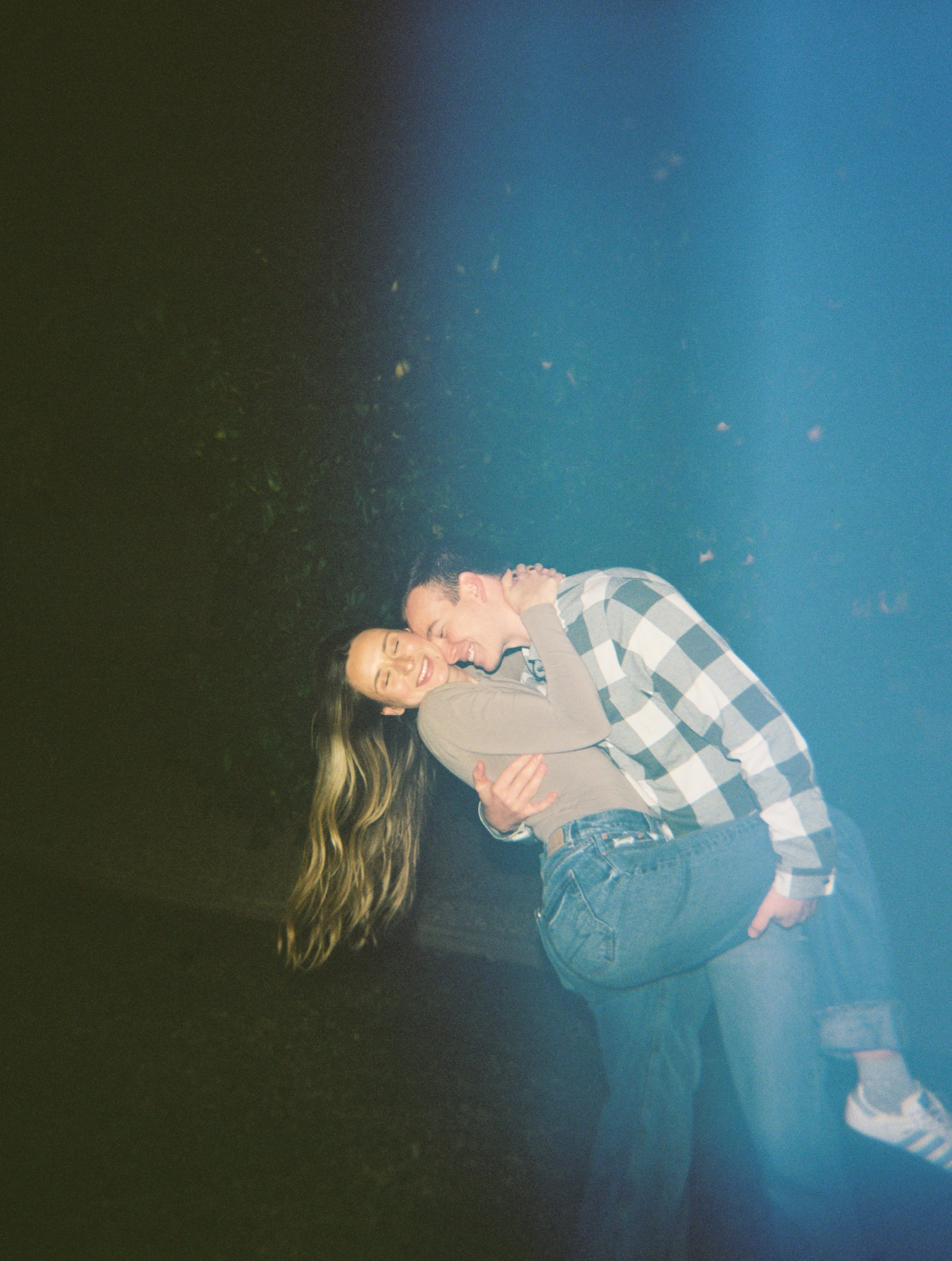 Two young women and a young man are smiling and hugging each other outdoors at night. The woman on the left has long blonde hair and is wearing a beige sweatshirt. The man on the right has short dark hair and is wearing a black and white checkered sh