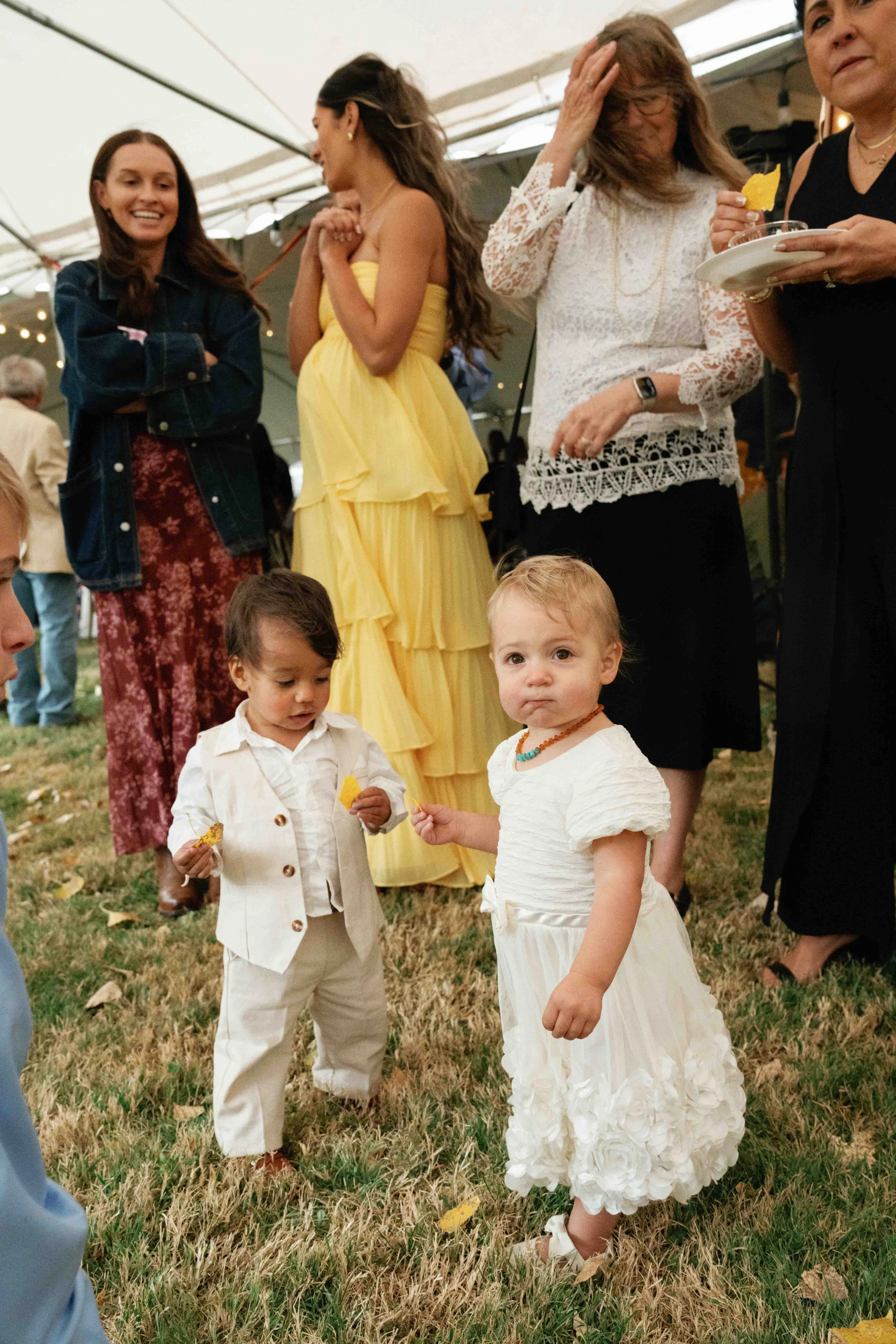Children and adults gathered outdoors under a tent, with children in fancy dresses and adults in casual and semi-formal attire, at a social event or celebration.