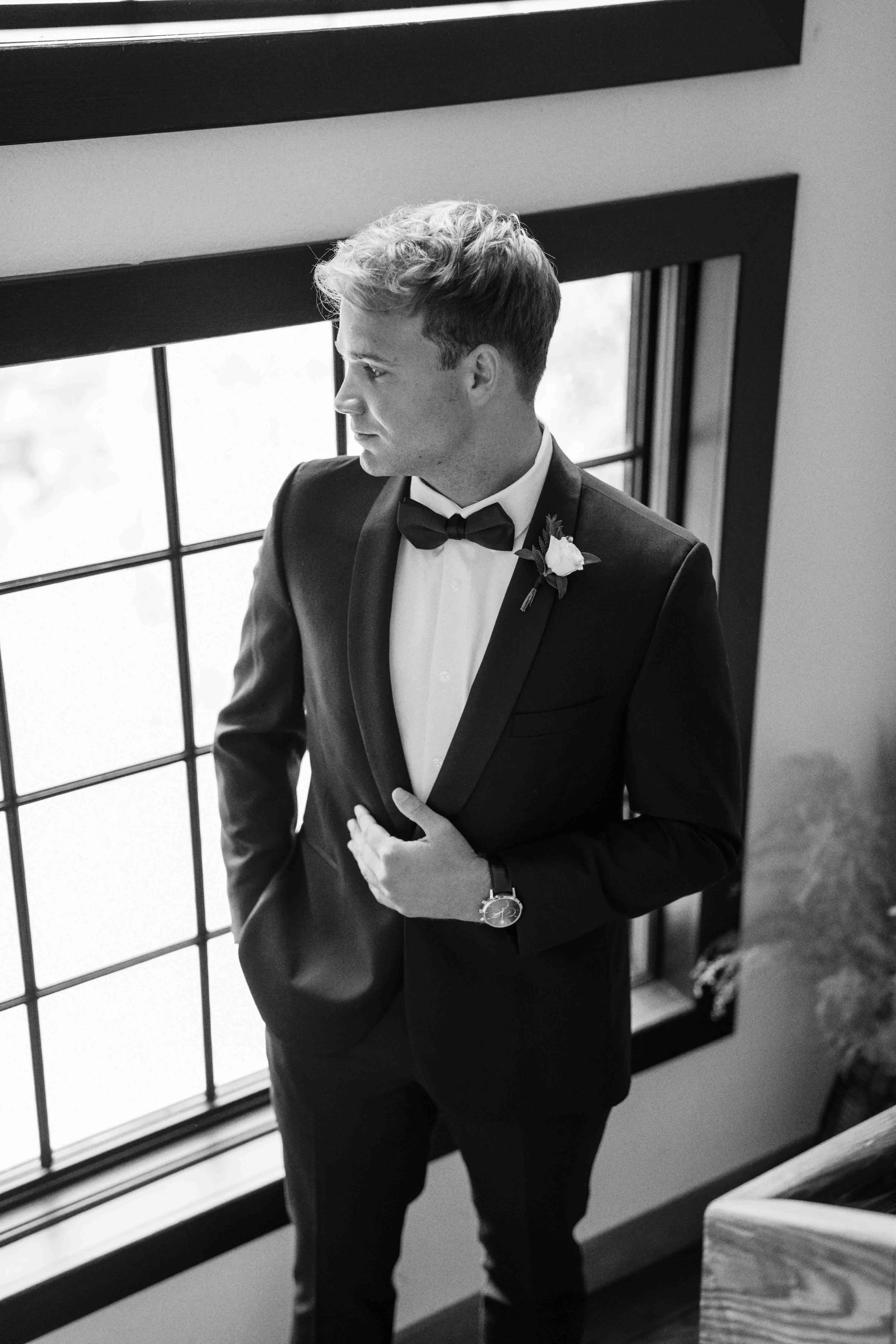 A young man in a tuxedo, wearing a bow tie, standing indoors near a large window with multiple panes, and looking to his left. He has a boutonniere on his lapel and a watch on his left wrist.