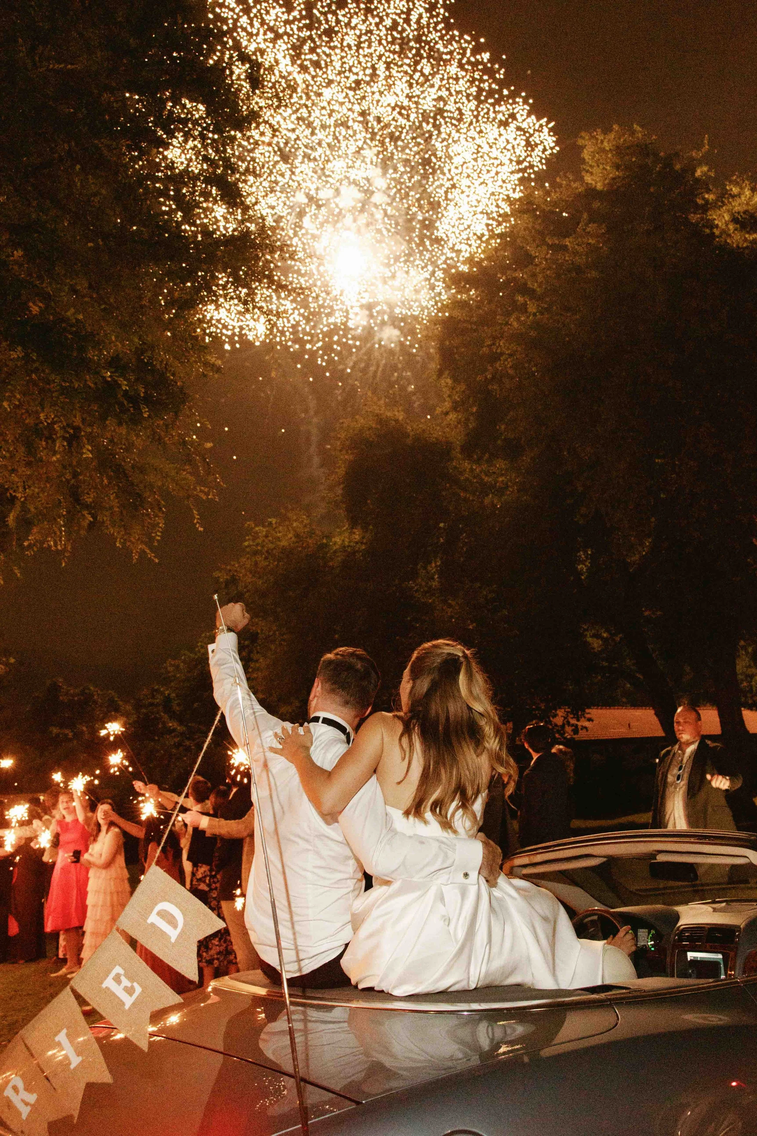 Couple dressed in wedding attire sitting on top of the car during nighttime celebration with sparklers, fireworks, and people in the background.
