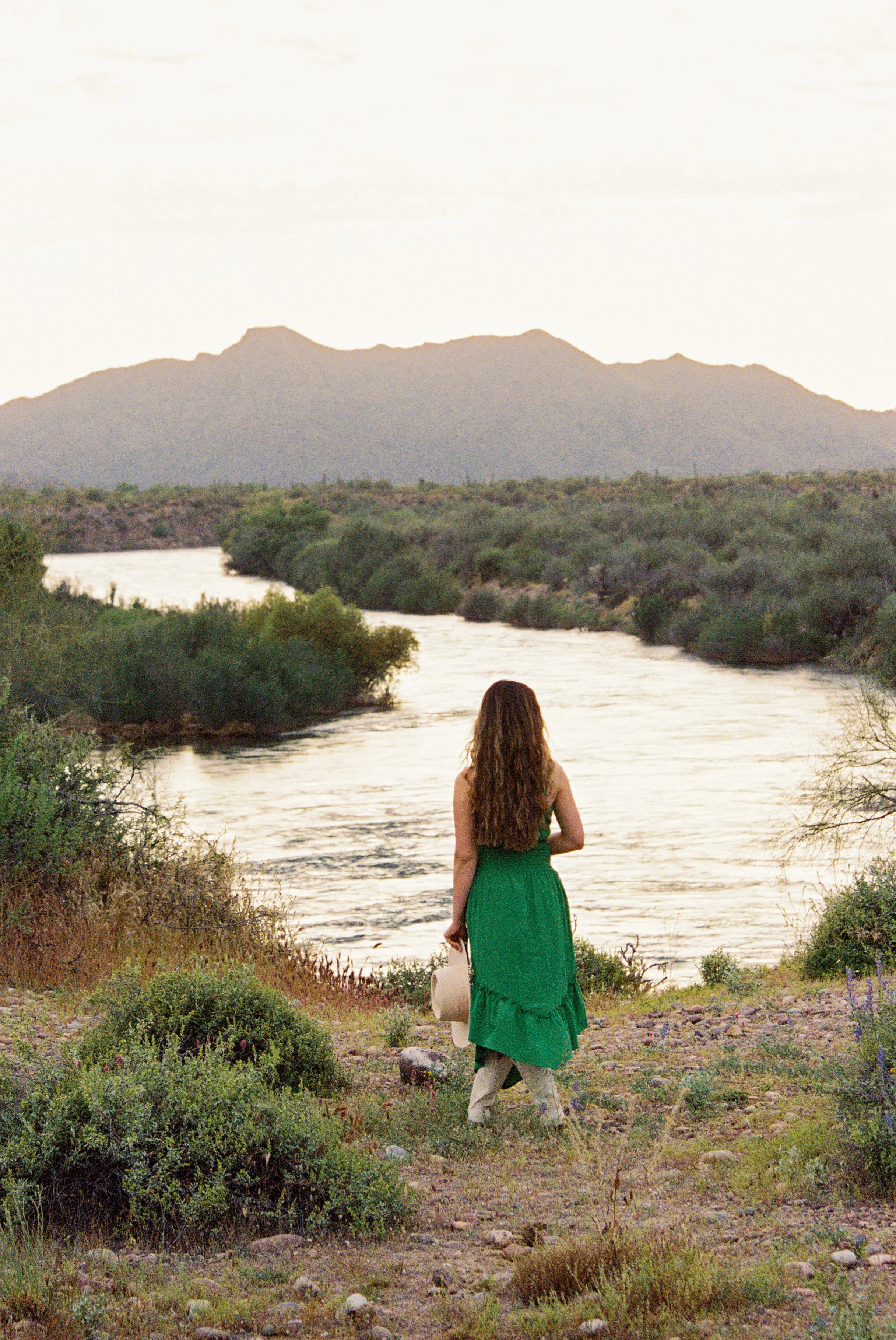 A woman with long, wavy brown hair in a green dress standing near a river, facing away, holding a beige hat and a white bag, with a mountain range in the background during a sunset or sunrise.