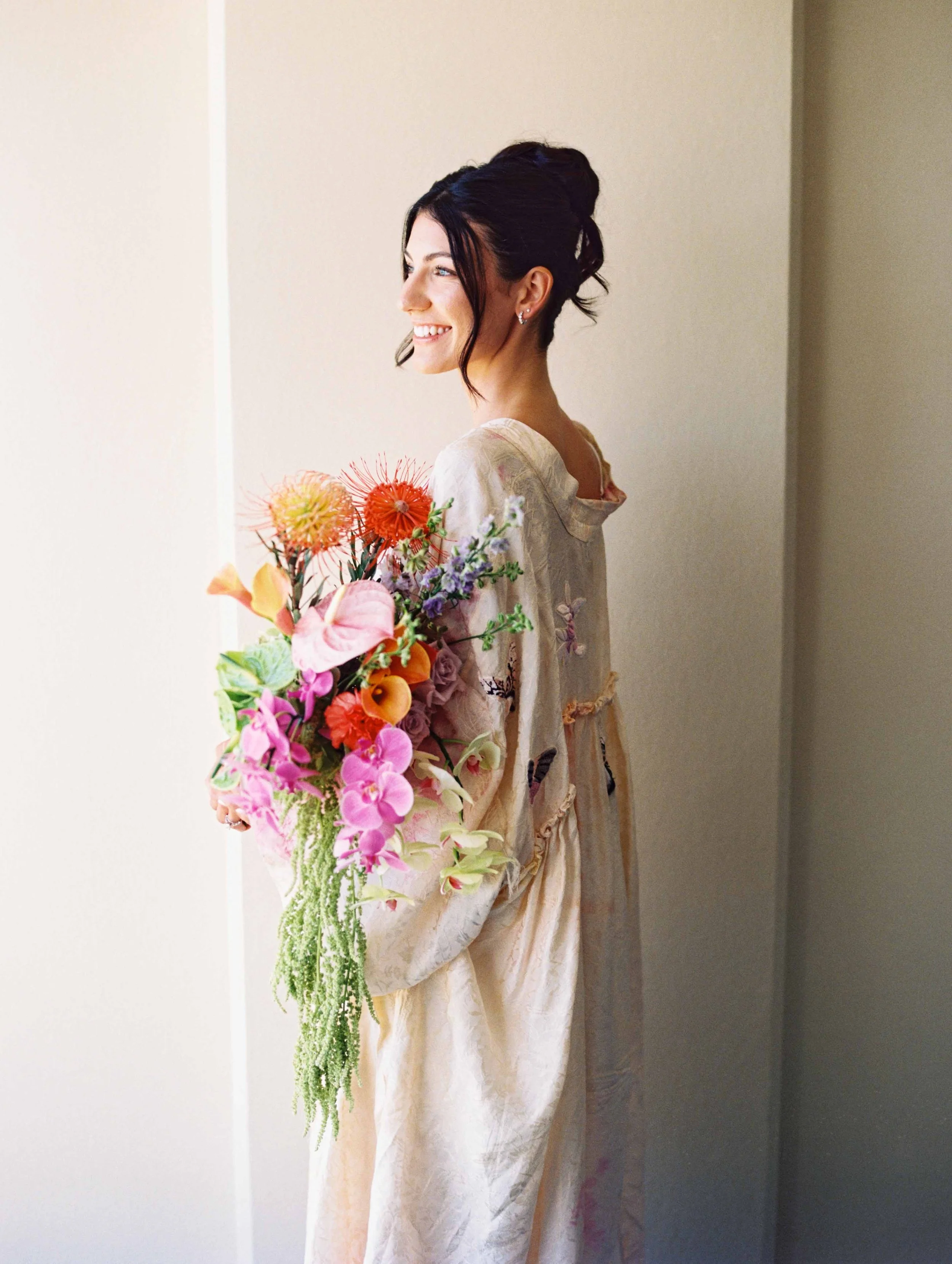 A woman in a cream-colored dress holding a large colorful bouquet of flowers, standing sideways against a plain wall, with dark hair styled in an updo and smiling.