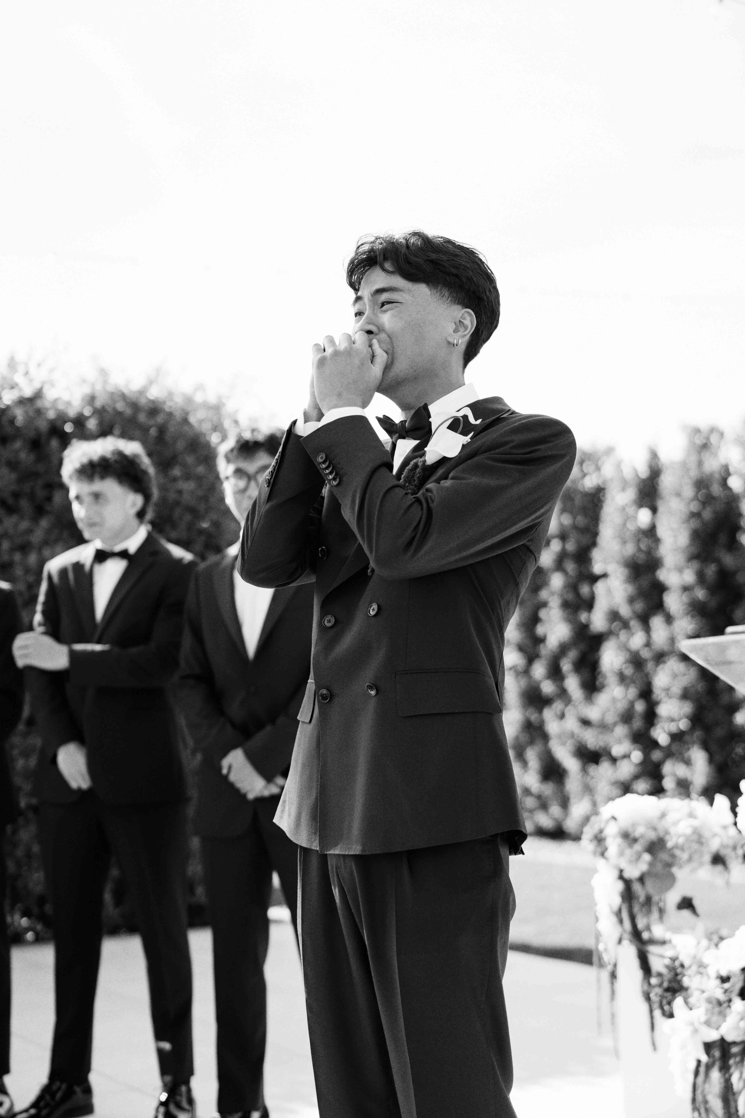A group of young men dressed in tuxedos standing outdoors with trees and flowers in the background, one of them covering his mouth with his hand and appearing to smile.