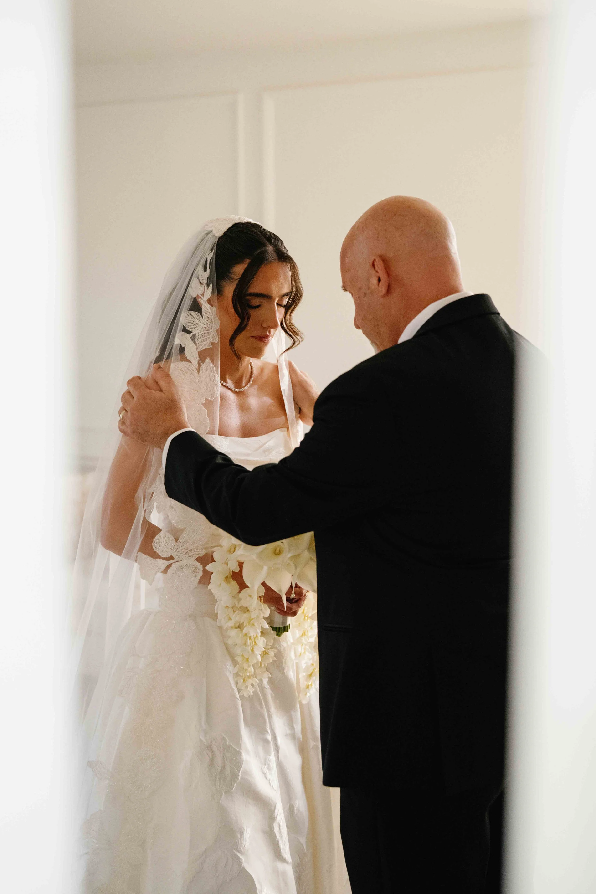 A bride and groom during a wedding ceremony, with the bride wearing a lace wedding gown and veil, holding a bouquet of white flowers, and the groom in a black suit, both with heads bowed and eyes closed.