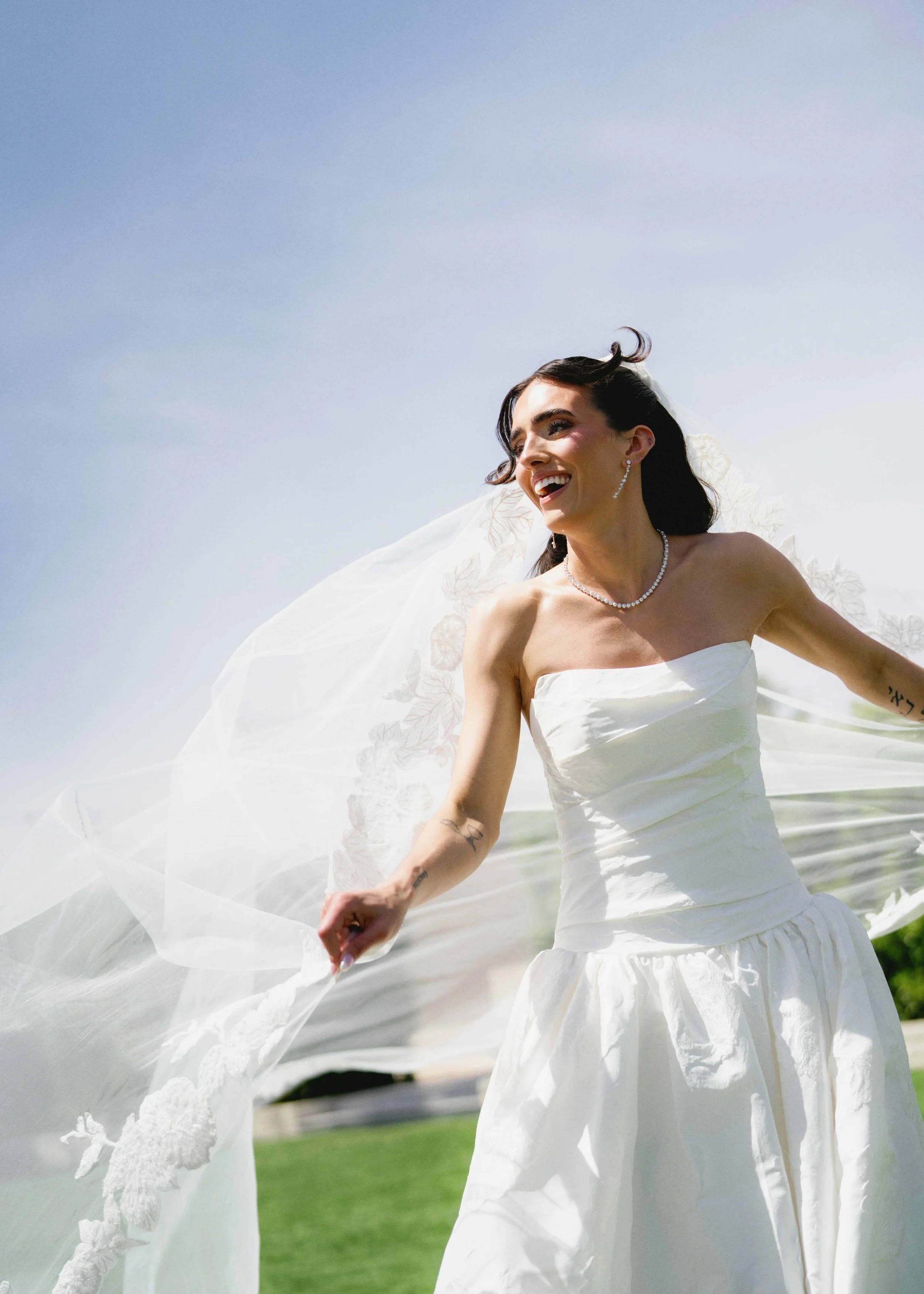 A woman in a wedding dress smiling outdoors, with a flowing shawl and jewelry, against a blue sky background.