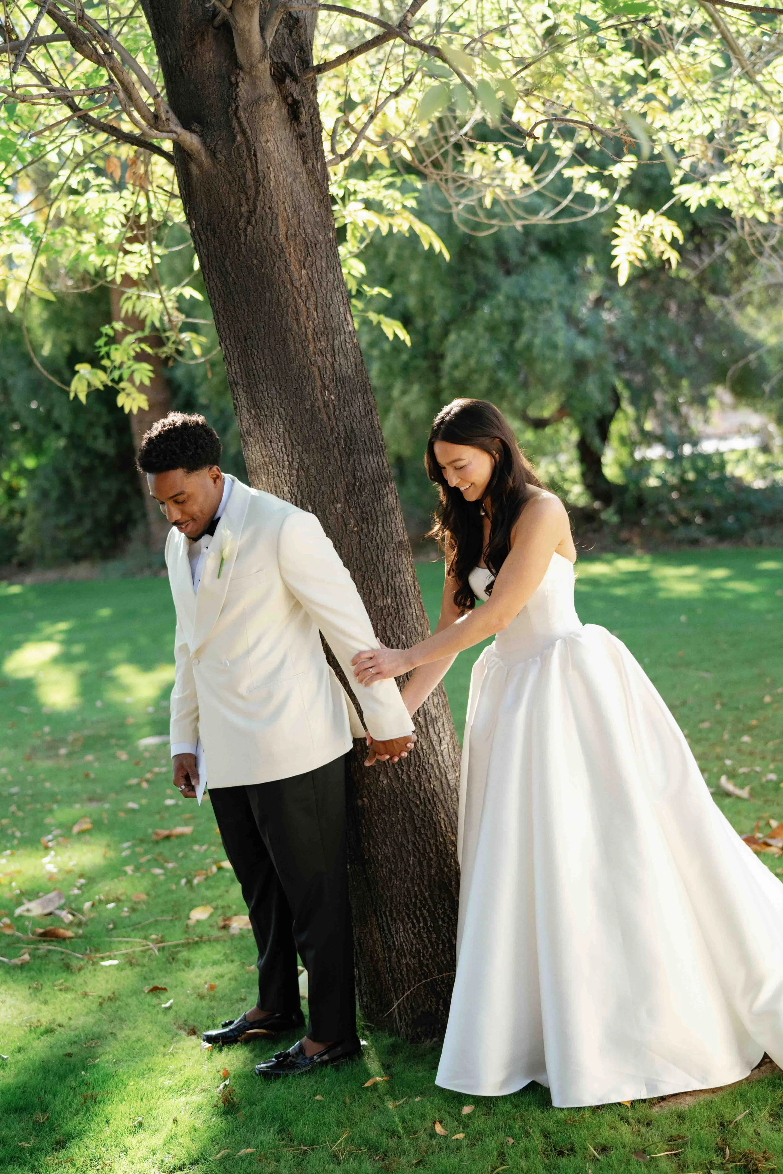A bride and groom holding hands and leaning against a tree in a park during their wedding. The groom is wearing a white tuxedo jacket, black pants, and black shoes. The bride is wearing a strapless white wedding gown with a full skirt and dark hair. 