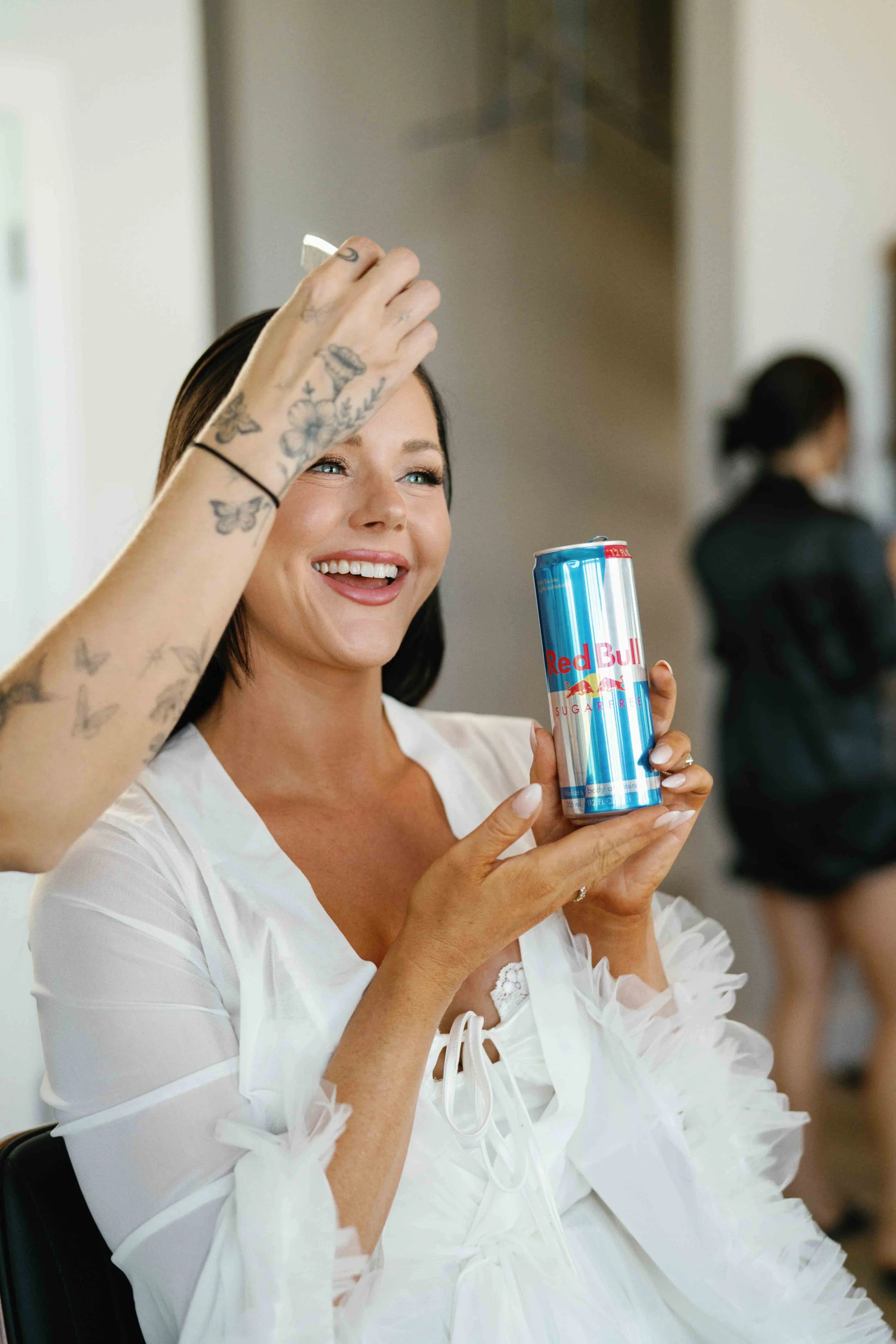 A woman with tattoos on her arms and dark hair smiling and holding a tall can of Red Bull energy drink, wearing a white dress with ruffle details, in a casual indoor setting.