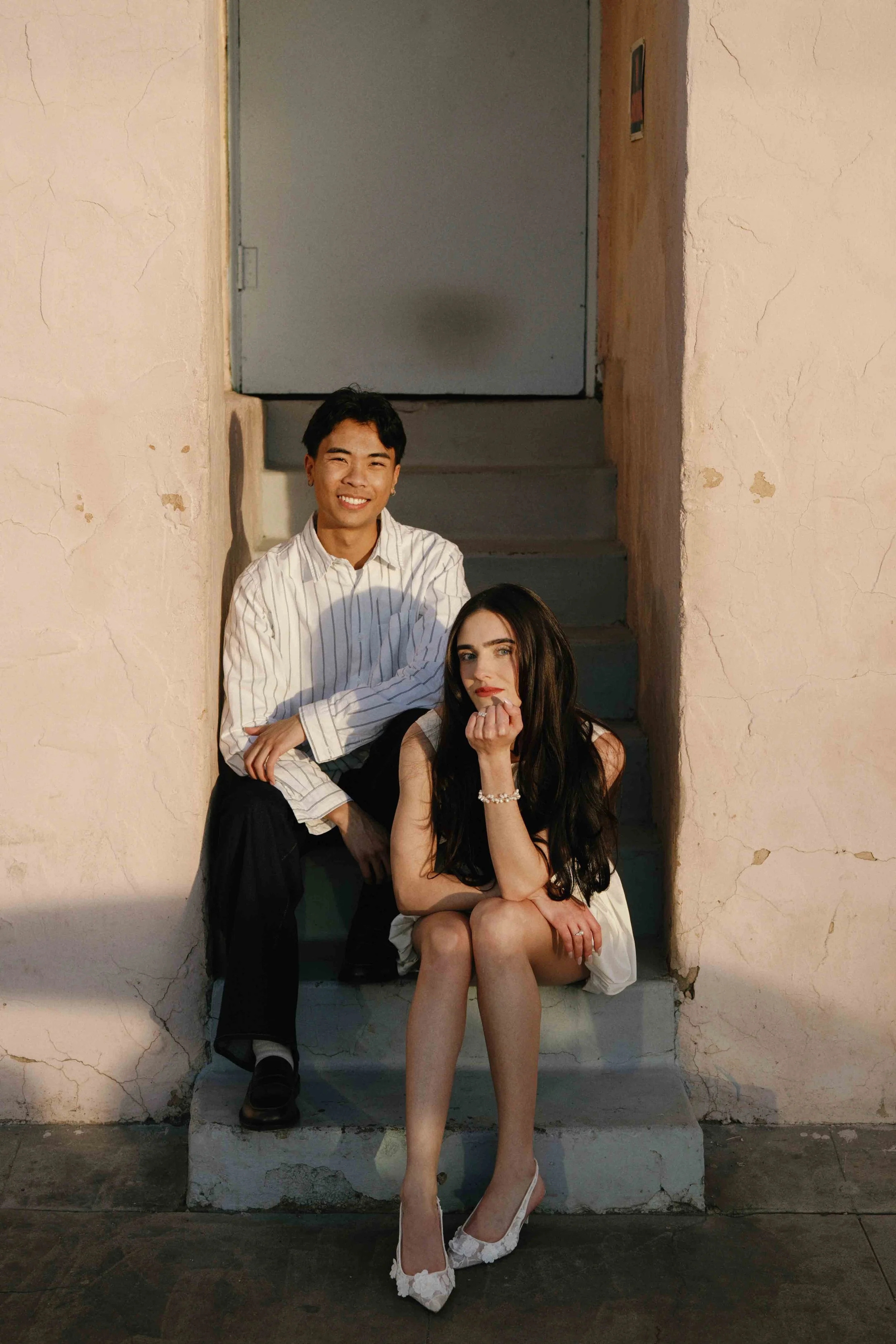 A young man with short dark hair and a smiling expression, wearing a white striped shirt, sitting on stairs. Next to him, a young woman with long dark hair and a serious expression, wearing a white dress and white high heels, sitting on the stairs wi