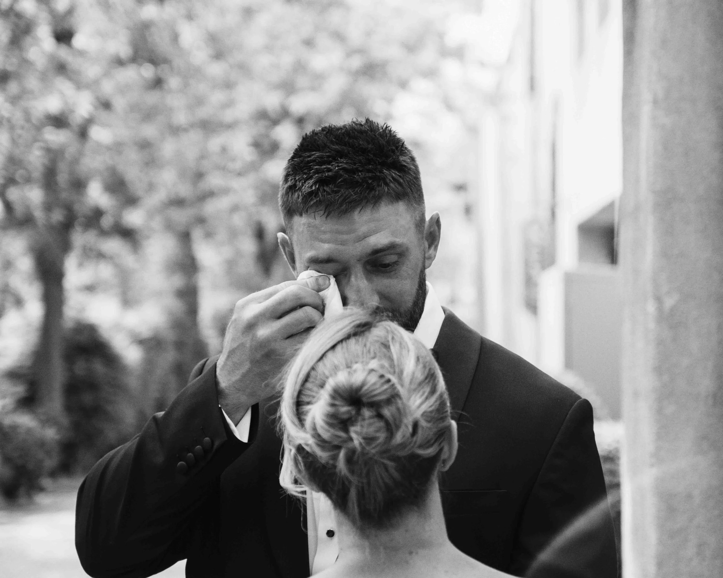 Black and white photo of a groom and bride, with the groom adjusting his tie, the bride below with her hair in a bun, and some guests in the background.