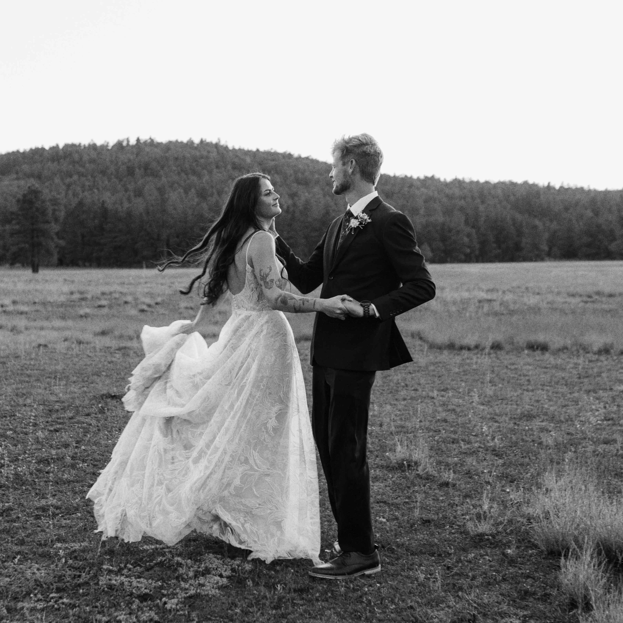 A black-and-white photo of a couple standing outdoors on grass, holding hands and facing each other, with a hilly landscape in the background.