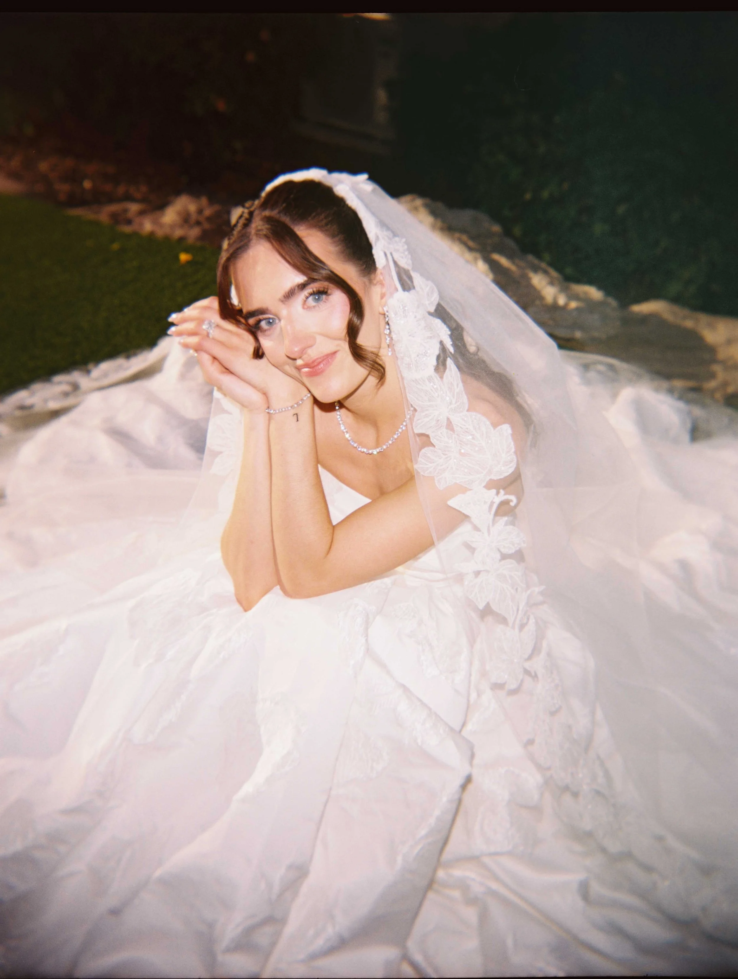 A woman lying on her side on a white wedding dress with lace details, wearing a pearl necklace and earrings, with shoulder-length brown hair and blue eyes, smiling gently at the camera at night or indoors.