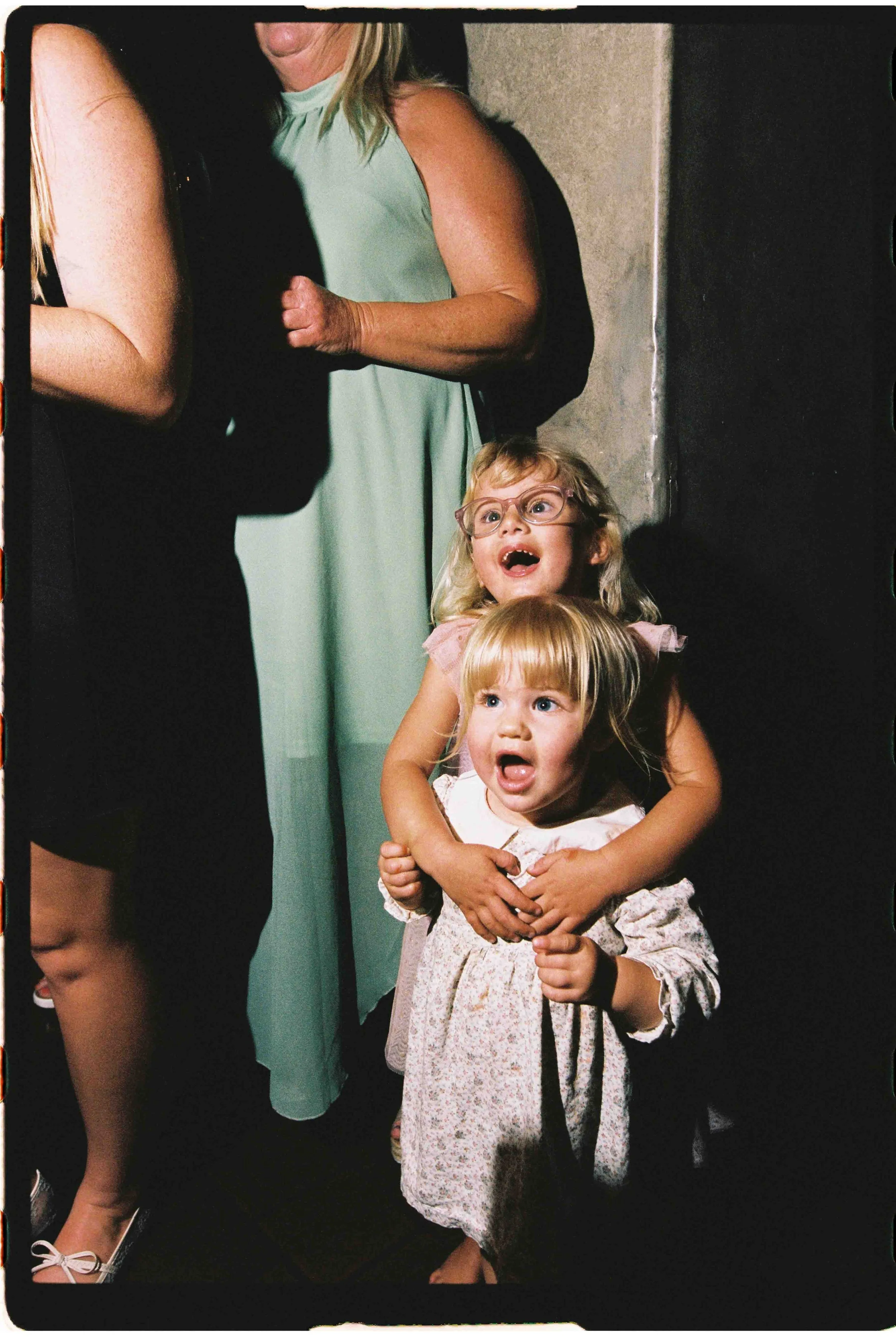 Two young girls, one with glasses, appear surprised and are being held gently by an adult woman. The scene is indoors with a concrete wall in the background.