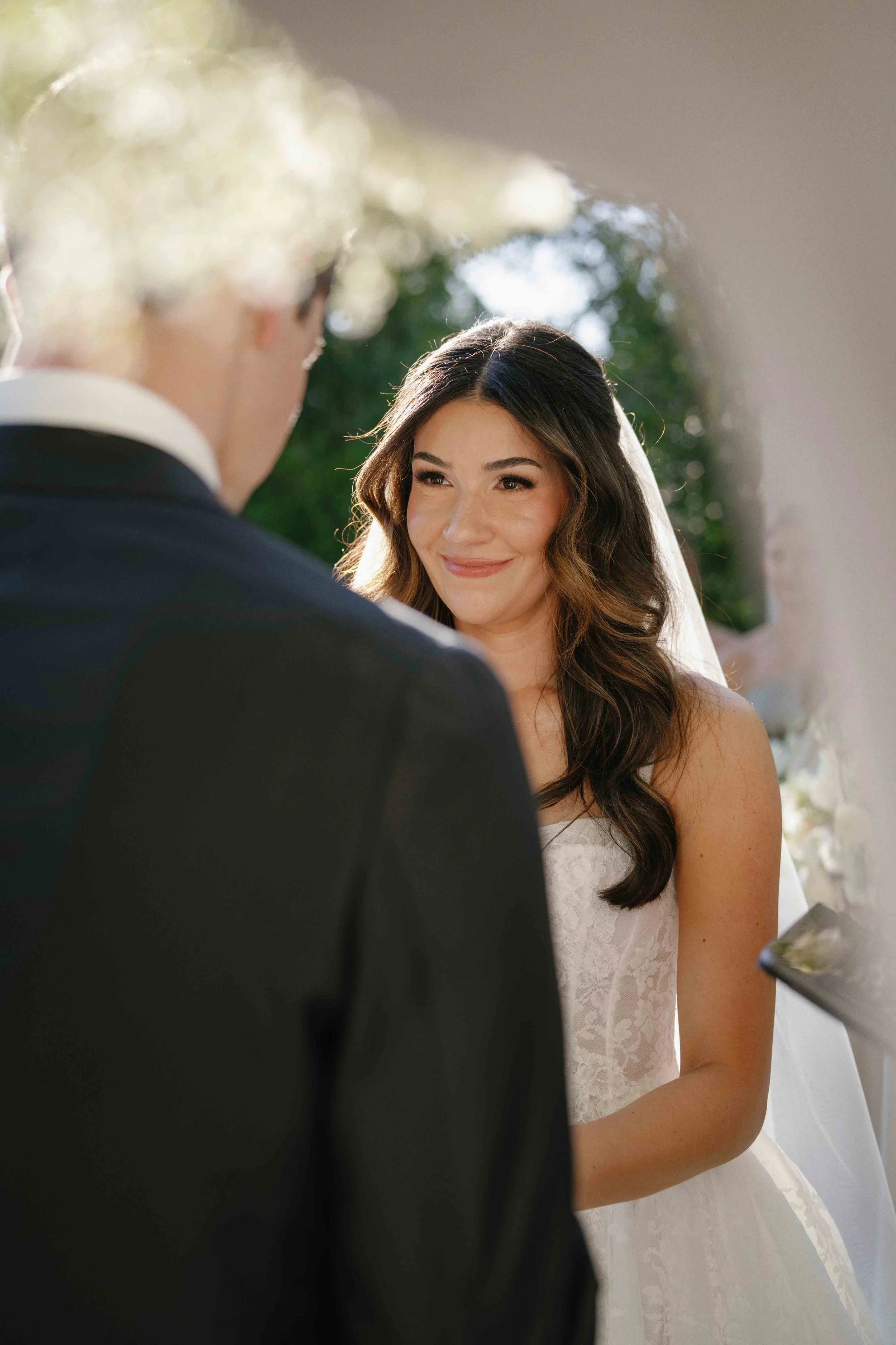 A bride with long, wavy brown hair, smiling and looking at a groom during their wedding ceremony.