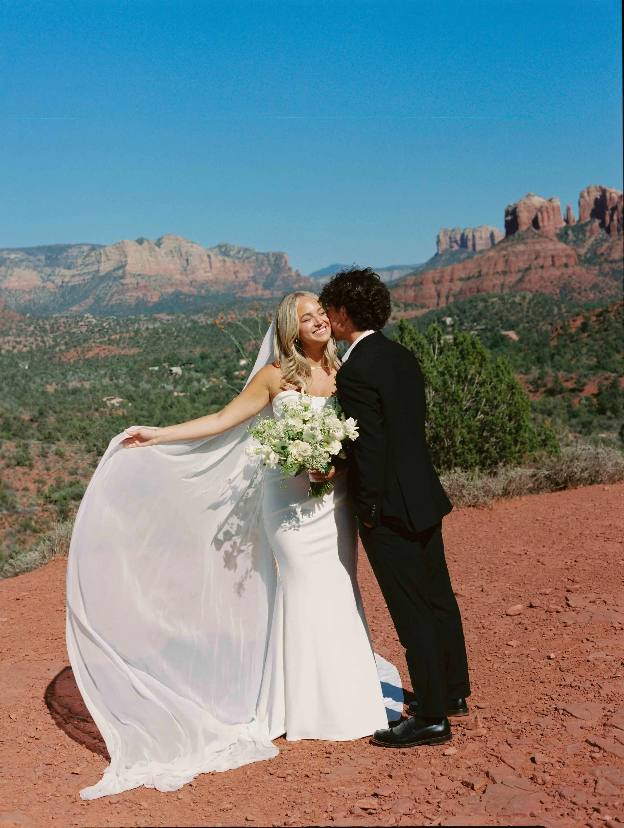 A bride and groom embrace in a scenic desert landscape with red rock formations and clear blue sky, the bride holding a bouquet and smiling, the groom dressed in a black suit.