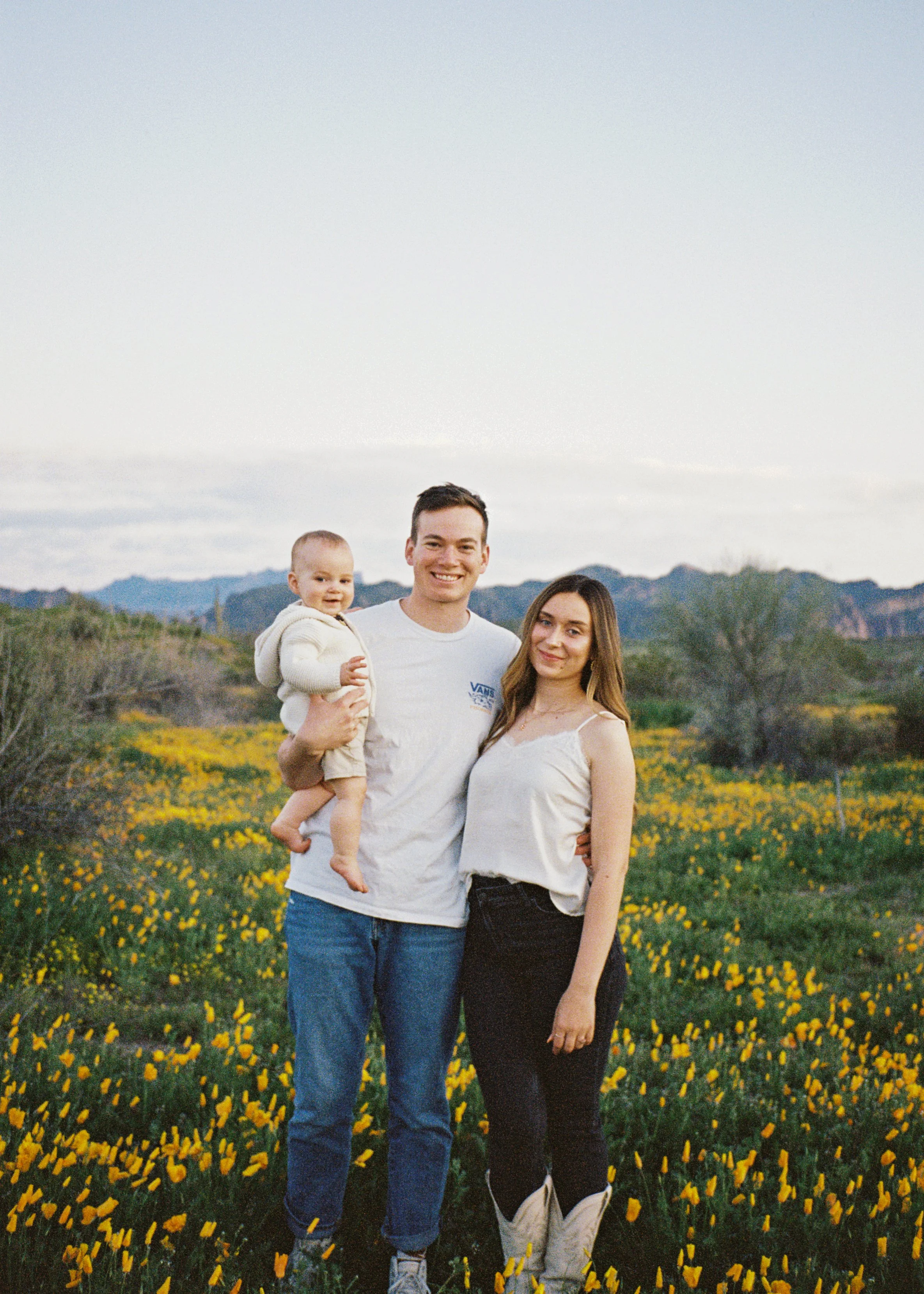 A young family of three, with a man, woman, and a child, standing close together in a field of yellow flowers during sunset, with mountains in the background.
