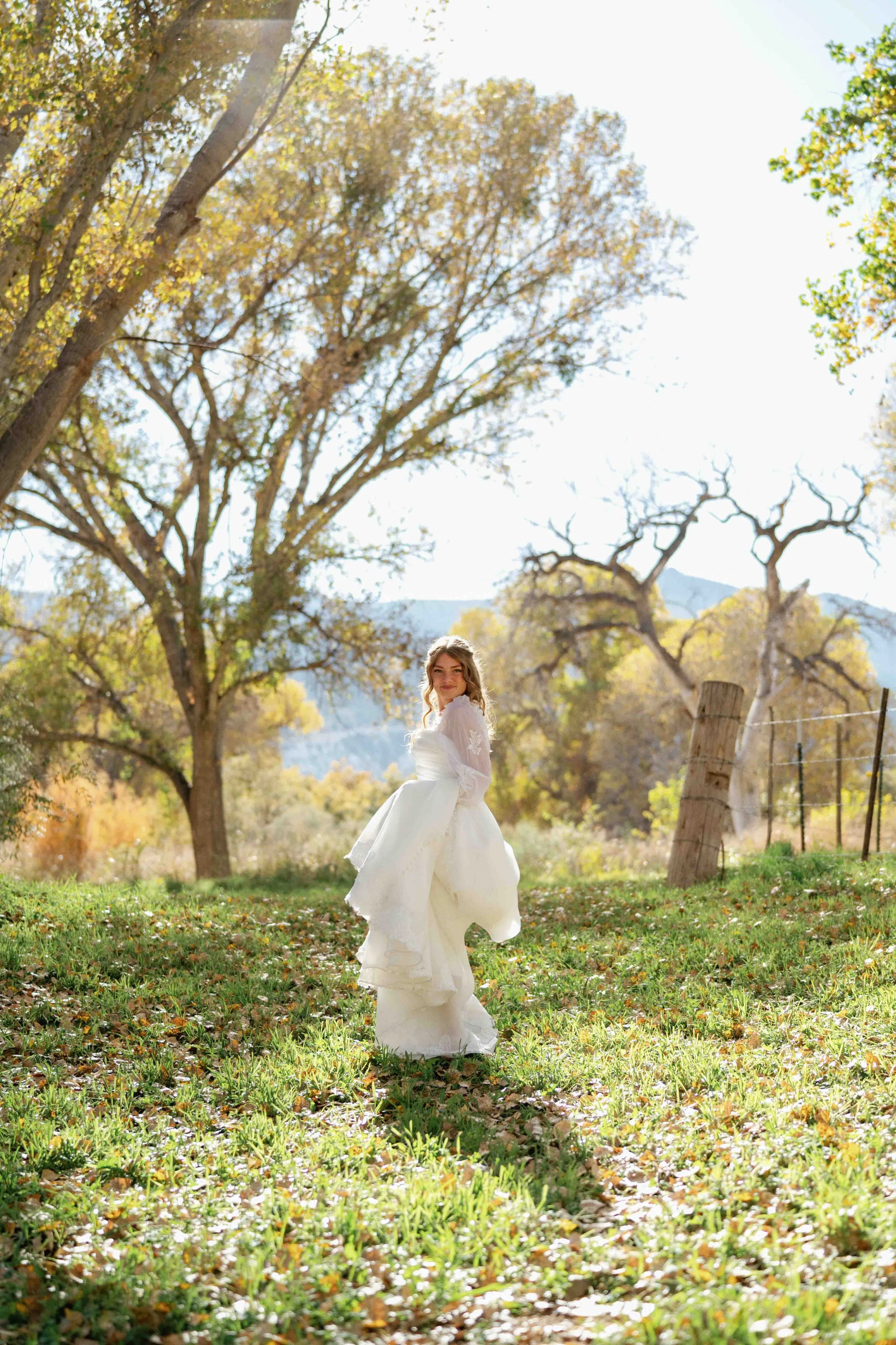 A woman in a white wedding dress standing outdoors under a clear sky, surrounded by trees with autumn leaves, grassy ground, and a wooden fence post.