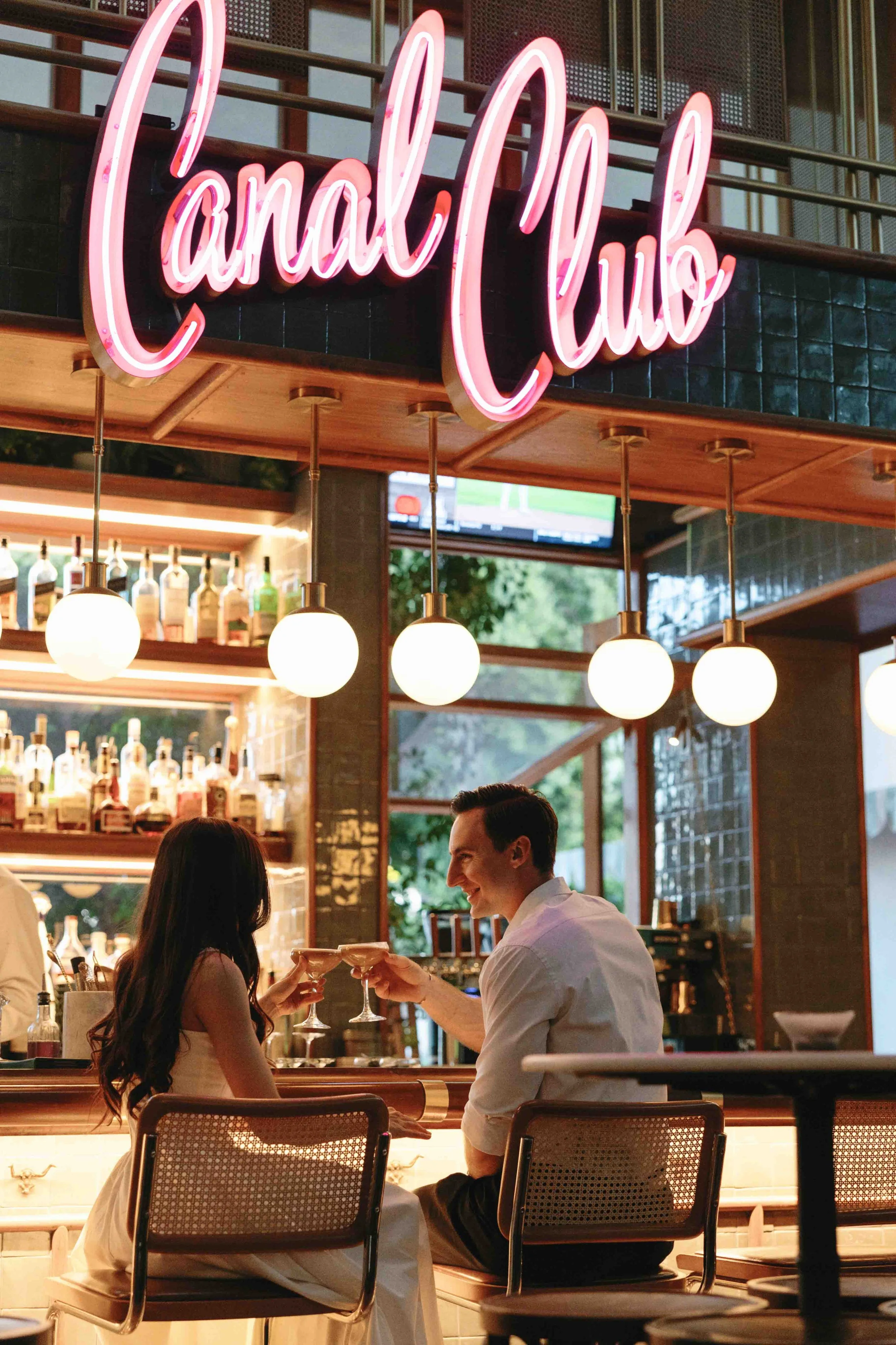 A romantic restaurant scene with a man and woman sharing a toast with cocktails, smiling at each other, in a warmly lit setting with a neon sign reading 'Local Loco' above them.