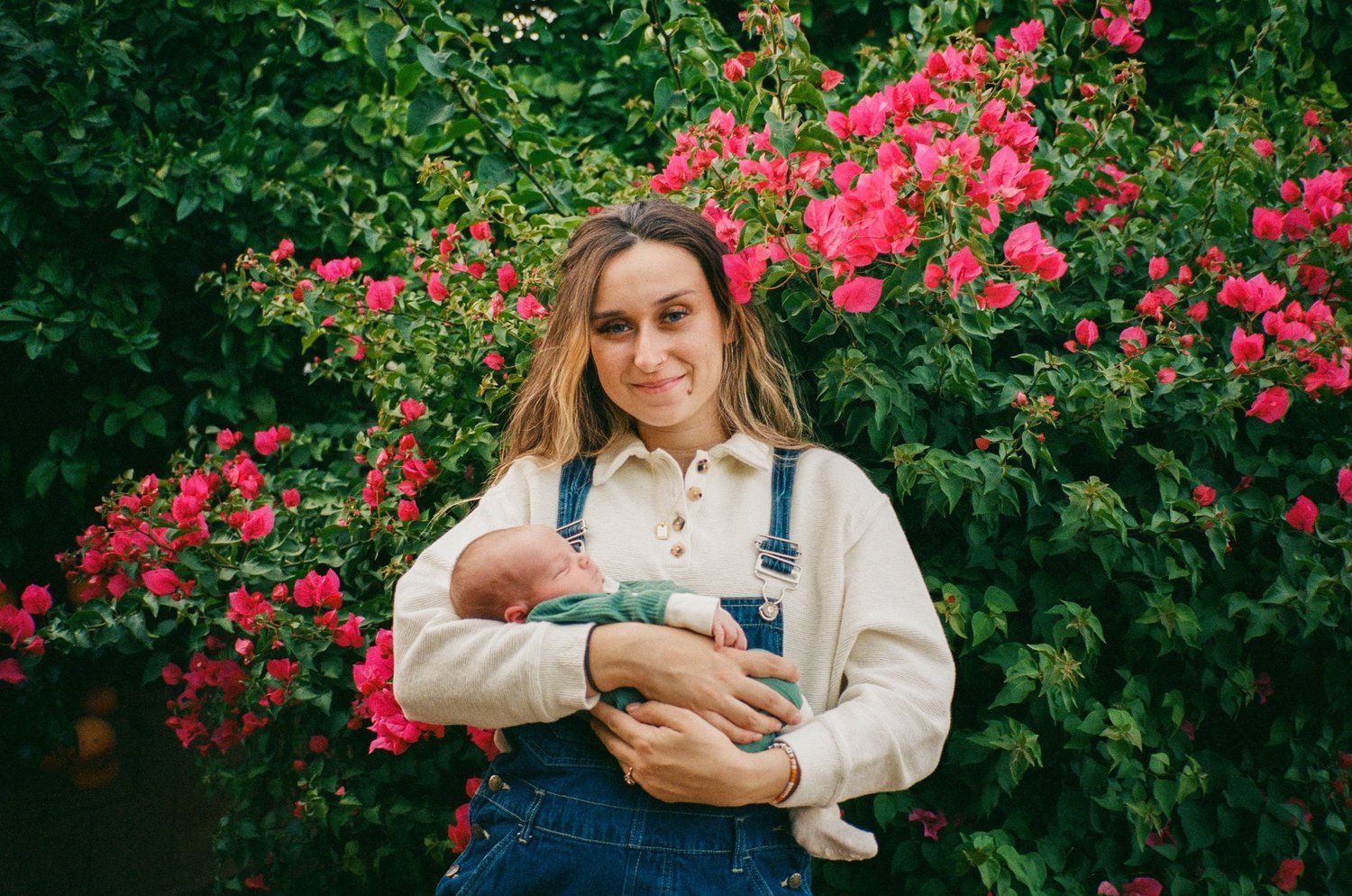 A young woman in a cream sweater and denim overalls holding a sleeping baby in front of a bush with bright pink flowers.