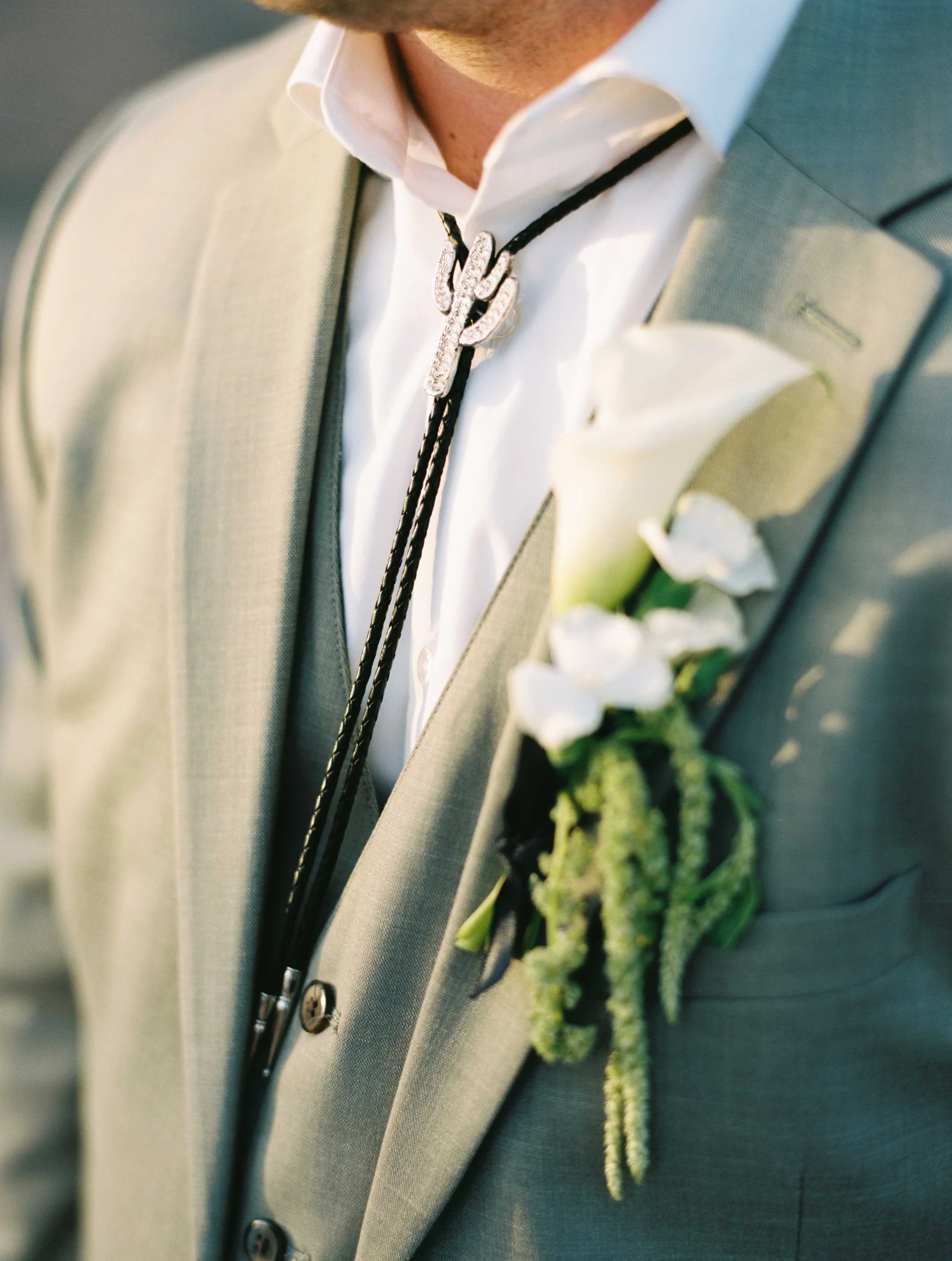 Close-up of a man wearing a light gray suit with a white shirt underneath. He has a black and silver necklace with a silver hand-shaped pendant and a green boutonniere pinned to his lapel.