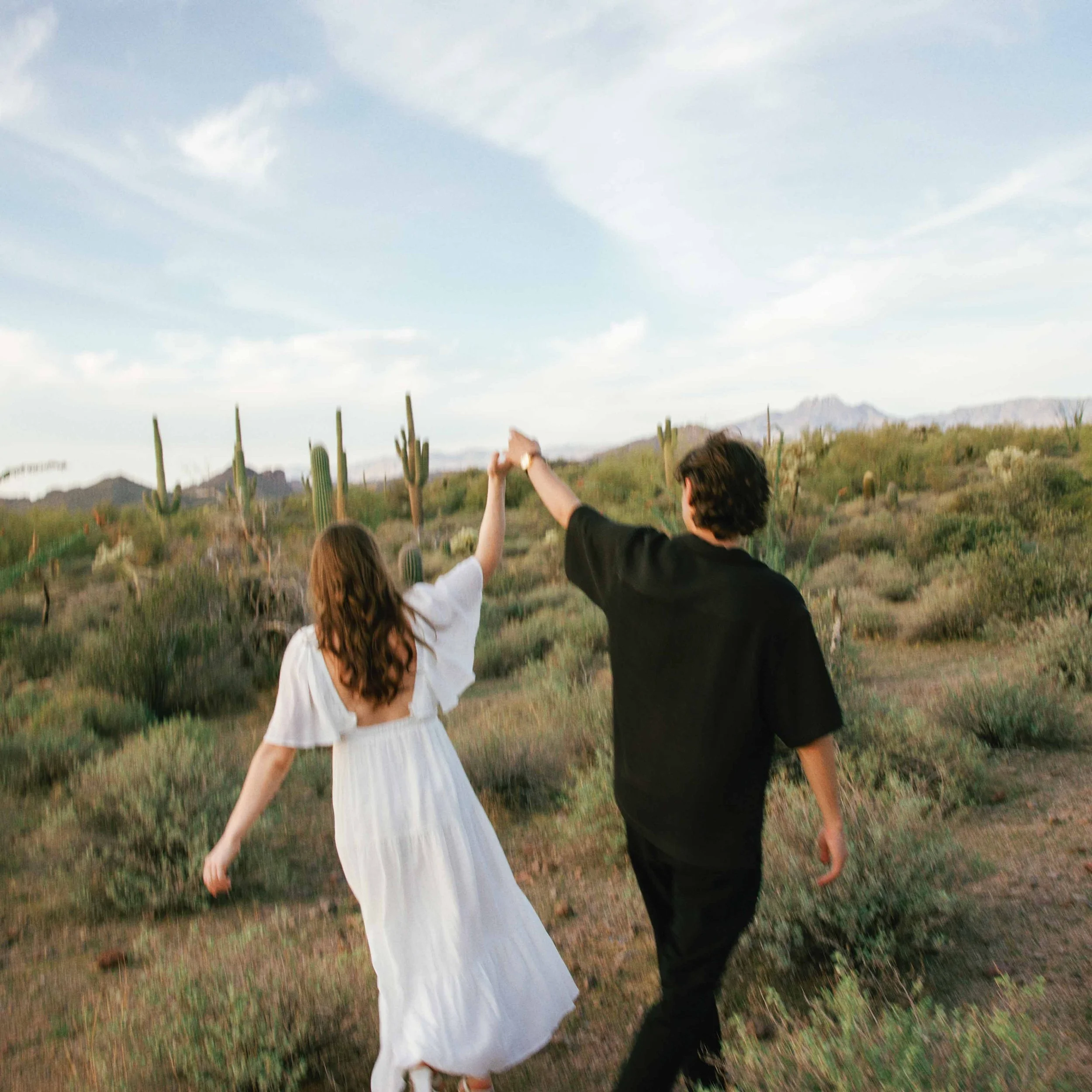 A young woman with long hair and a young man with short hair, holding hands while walking together in a desert landscape with cacti and mountains under a cloudy sky.