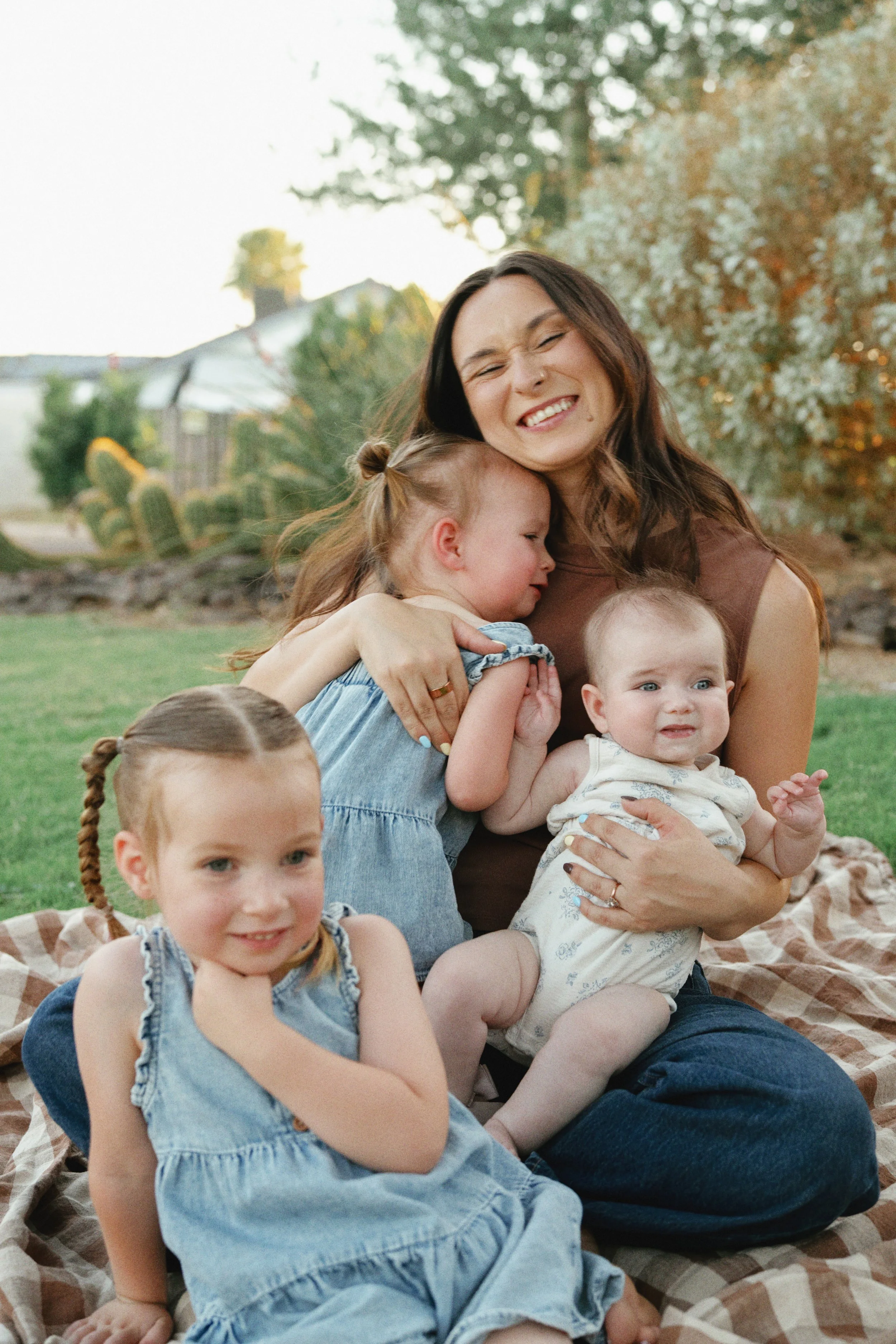 A woman with long brown hair and a delighted expression sits outdoors on a blanket, hugging and playing with several young children during what appears to be a family gathering or picnic in a garden or park setting, with trees and a house in the back