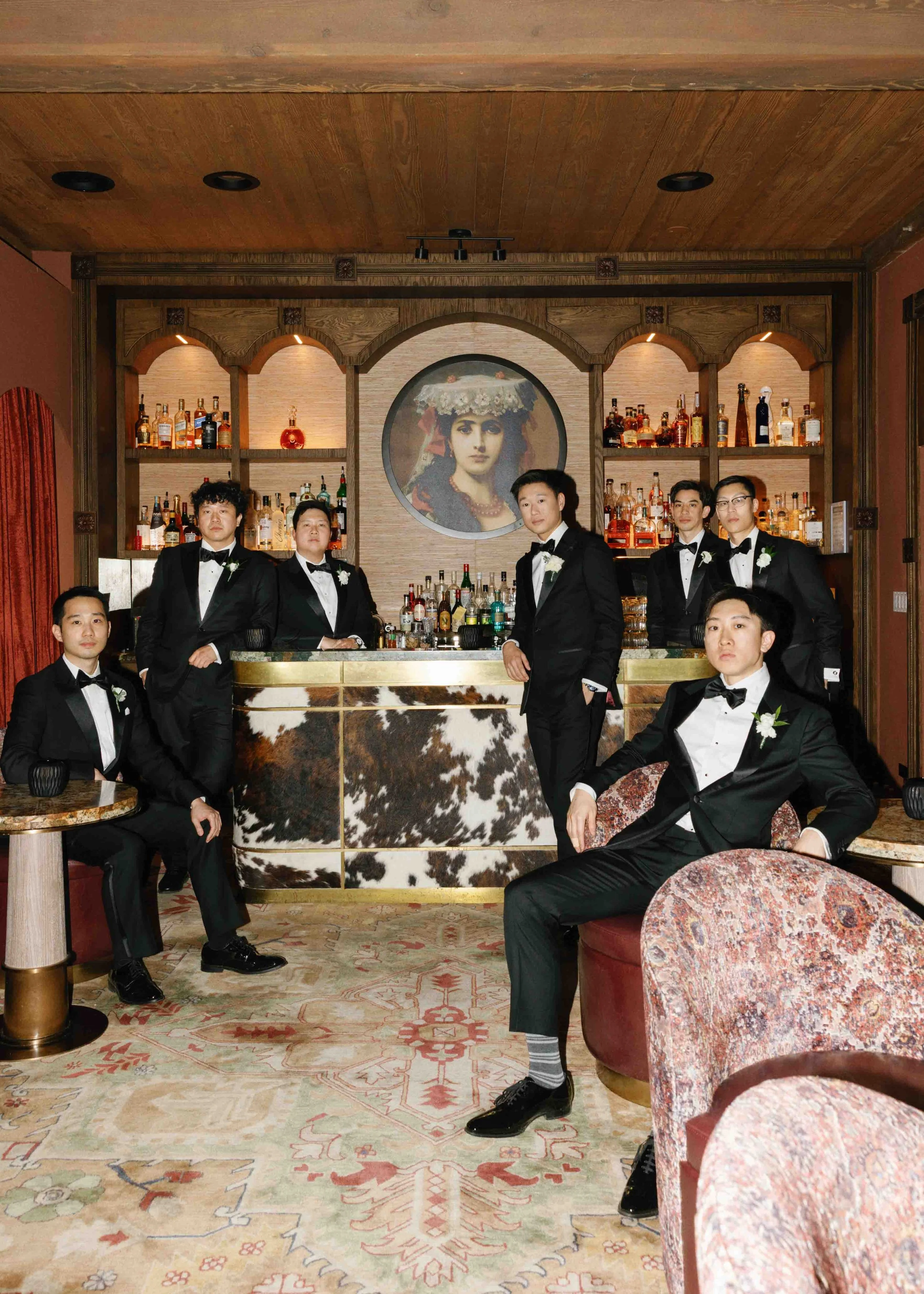 Group of seven men in tuxedos at a bar with a wooden background and a painting of a woman in a floral headdress.