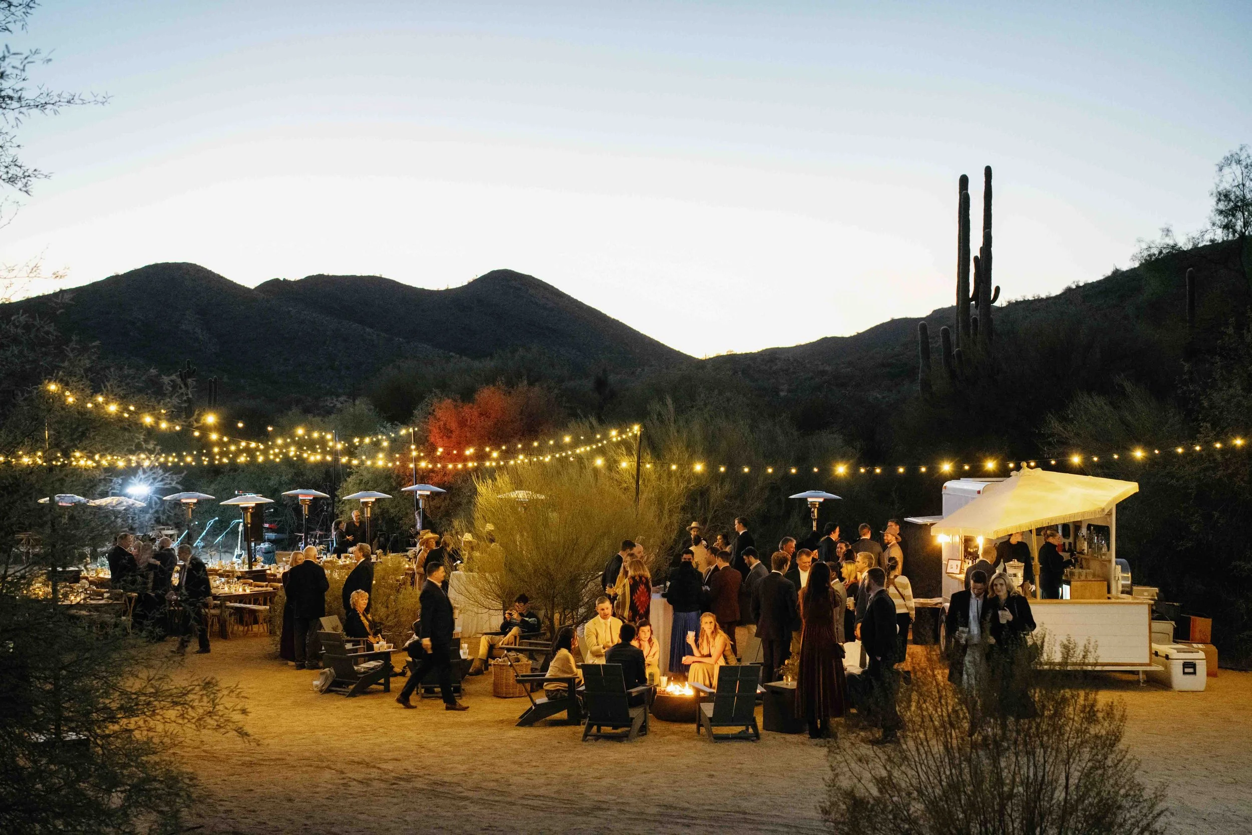 People gathered at an outdoor evening event with string lights, mountains, and saguaro cacti in the background.