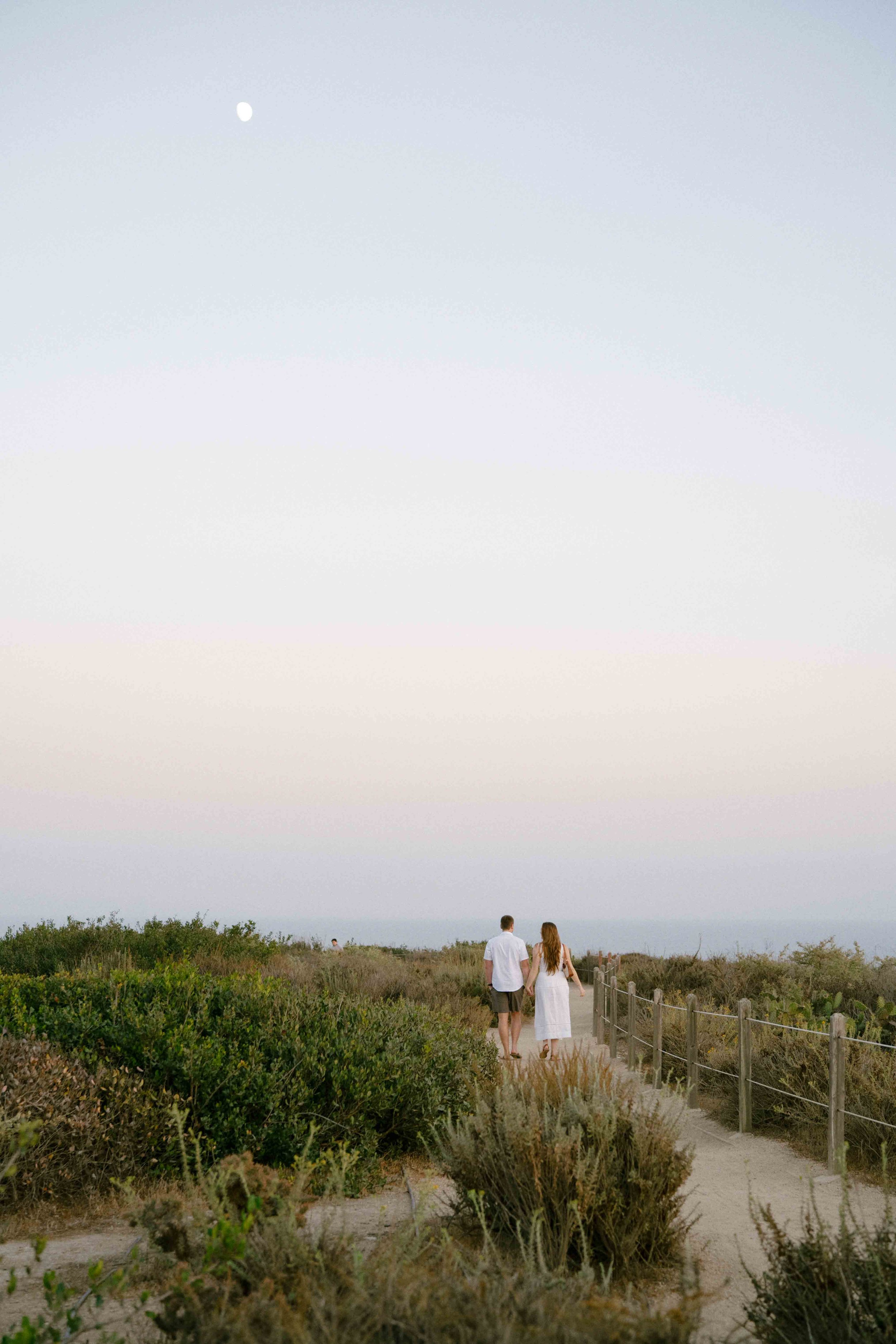 A couple walks along a sandy path surrounded by bushes and vegetation, under a pale sky with a faint moon visible in the distance.