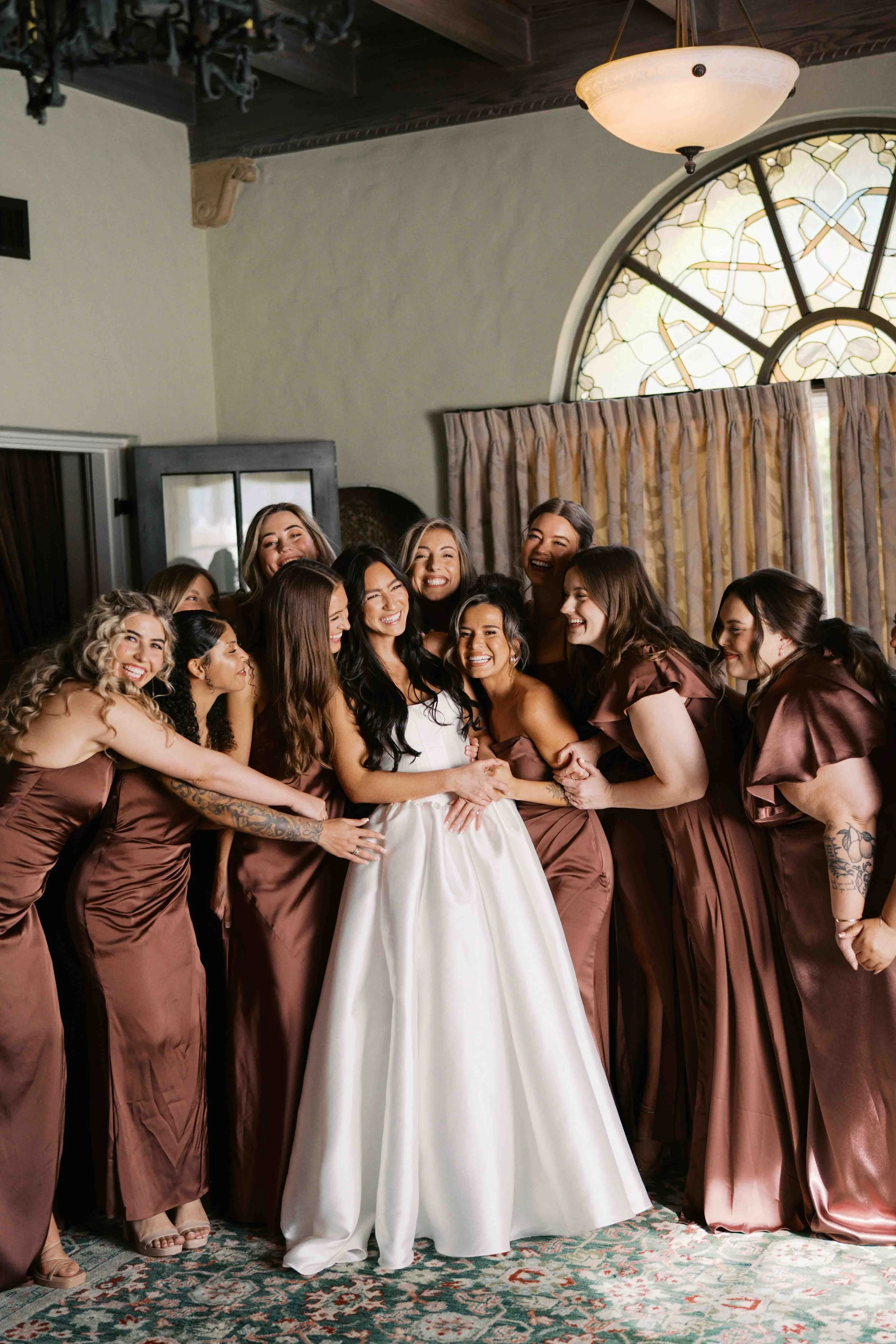 A group of women in matching brown bridesmaid dresses smiling and surrounding a bride in a white gown inside a room with a large stained glass window, ornate ceiling, and patterned carpet.