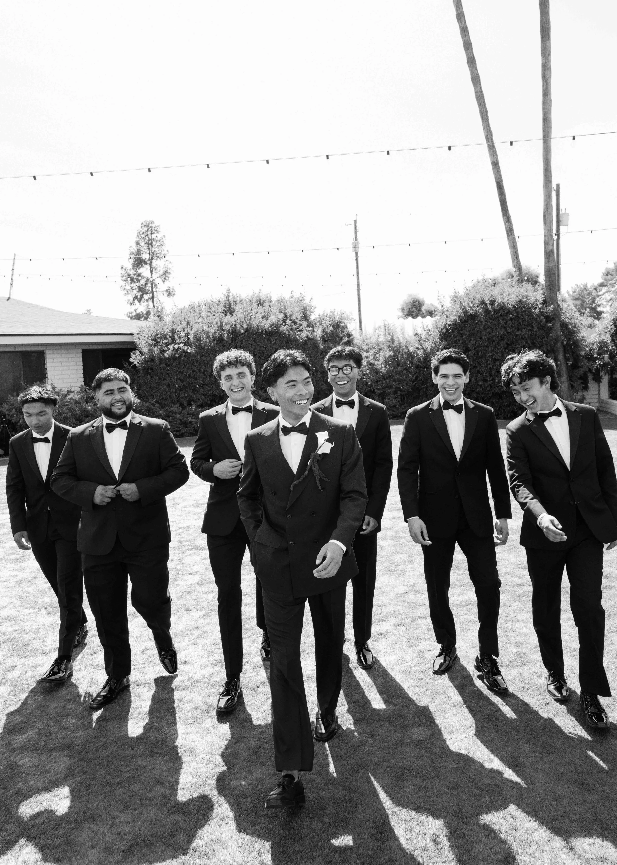 A group of young men in tuxedos walking in a line outdoors, casting long shadows on the grass, with trees and power lines in the background.