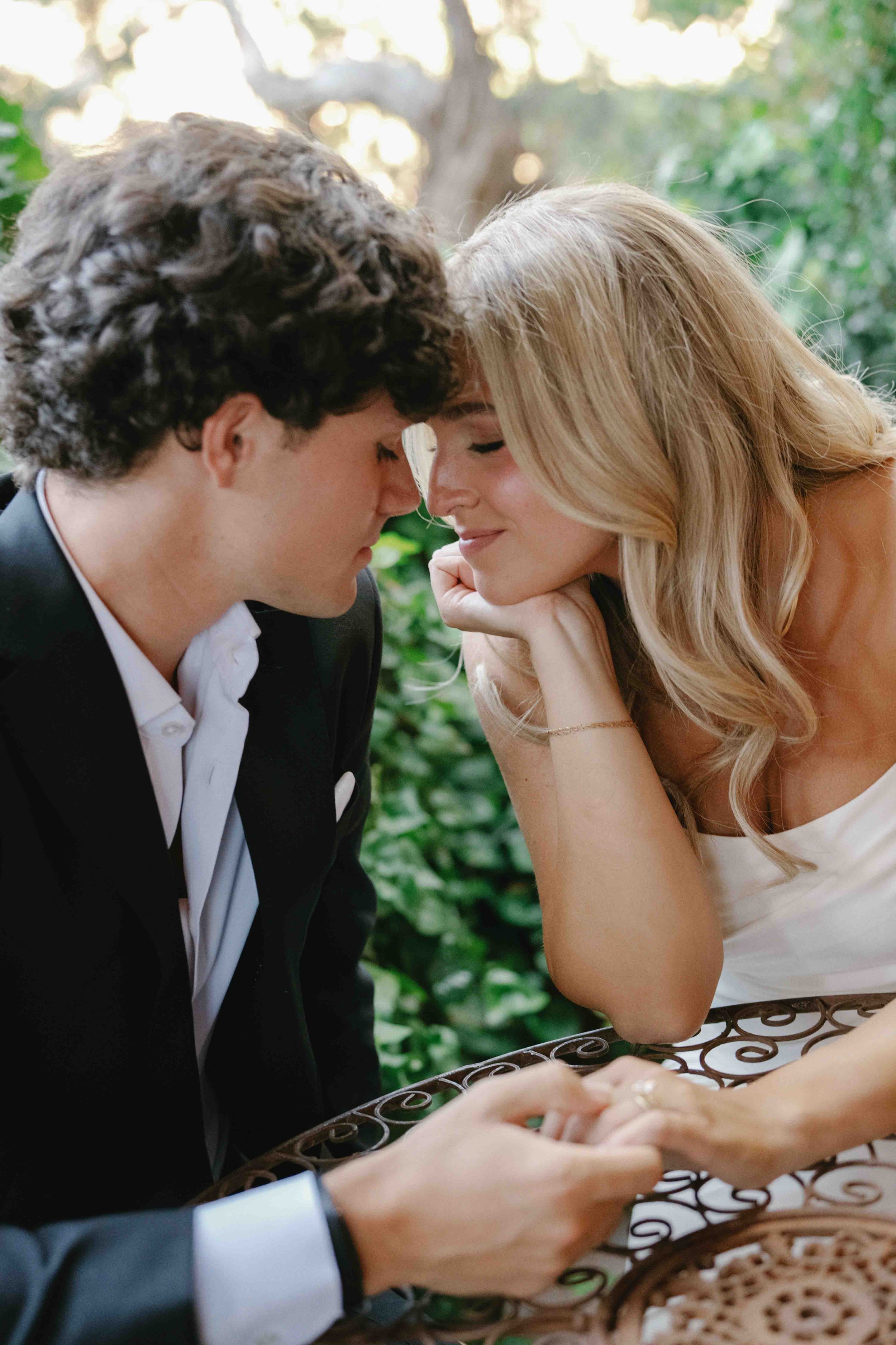 A couple with their foreheads touching, sitting close together at an outdoor table. The woman is smiling softly with her eyes closed, and the man is facing her with his eyes closed. The background is filled with green foliage and sunlight filtering t