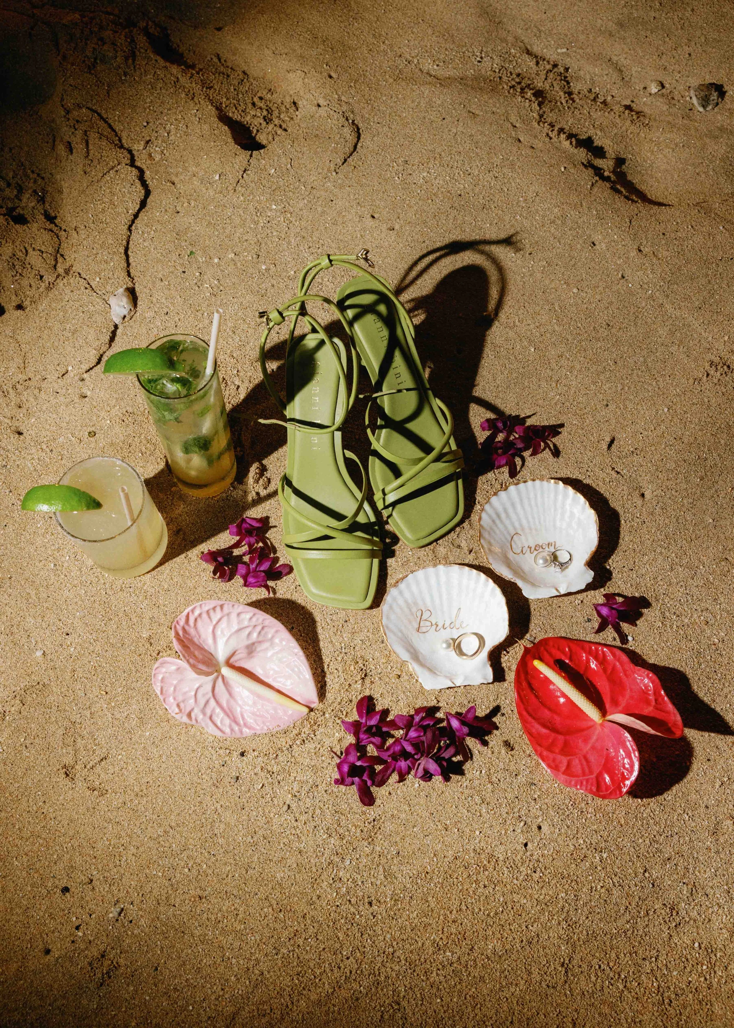 Green flip-flops, two glasses of drinks with lime wedges, seashells labeled 'Groom' and 'Bride' with rings, purple flowers, and red anthuriums on sand, suggesting a beach wedding setup.