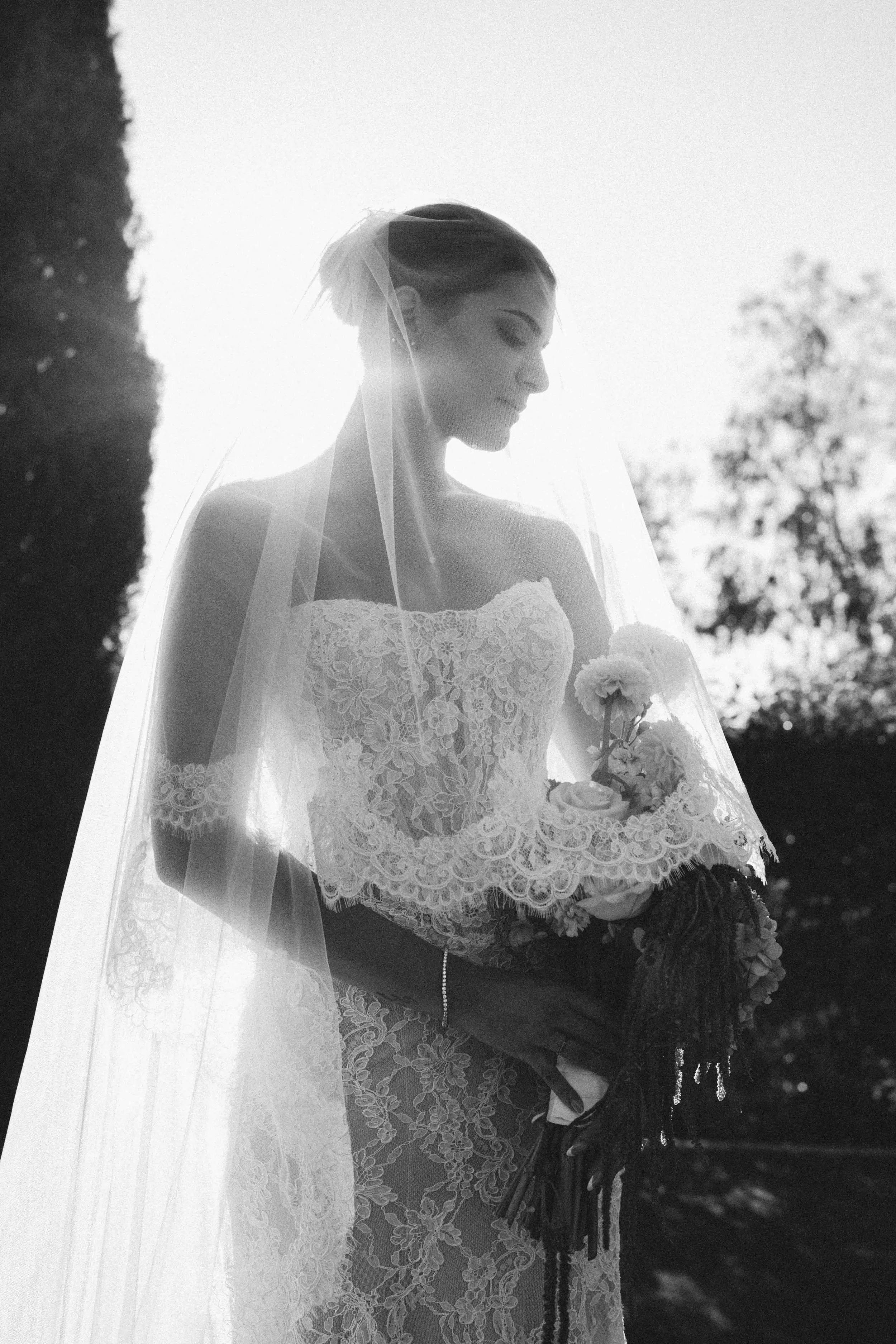 Black and white photograph of a bride in a lace wedding gown and veil, holding a bouquet of flowers, standing outdoors with trees in the background.