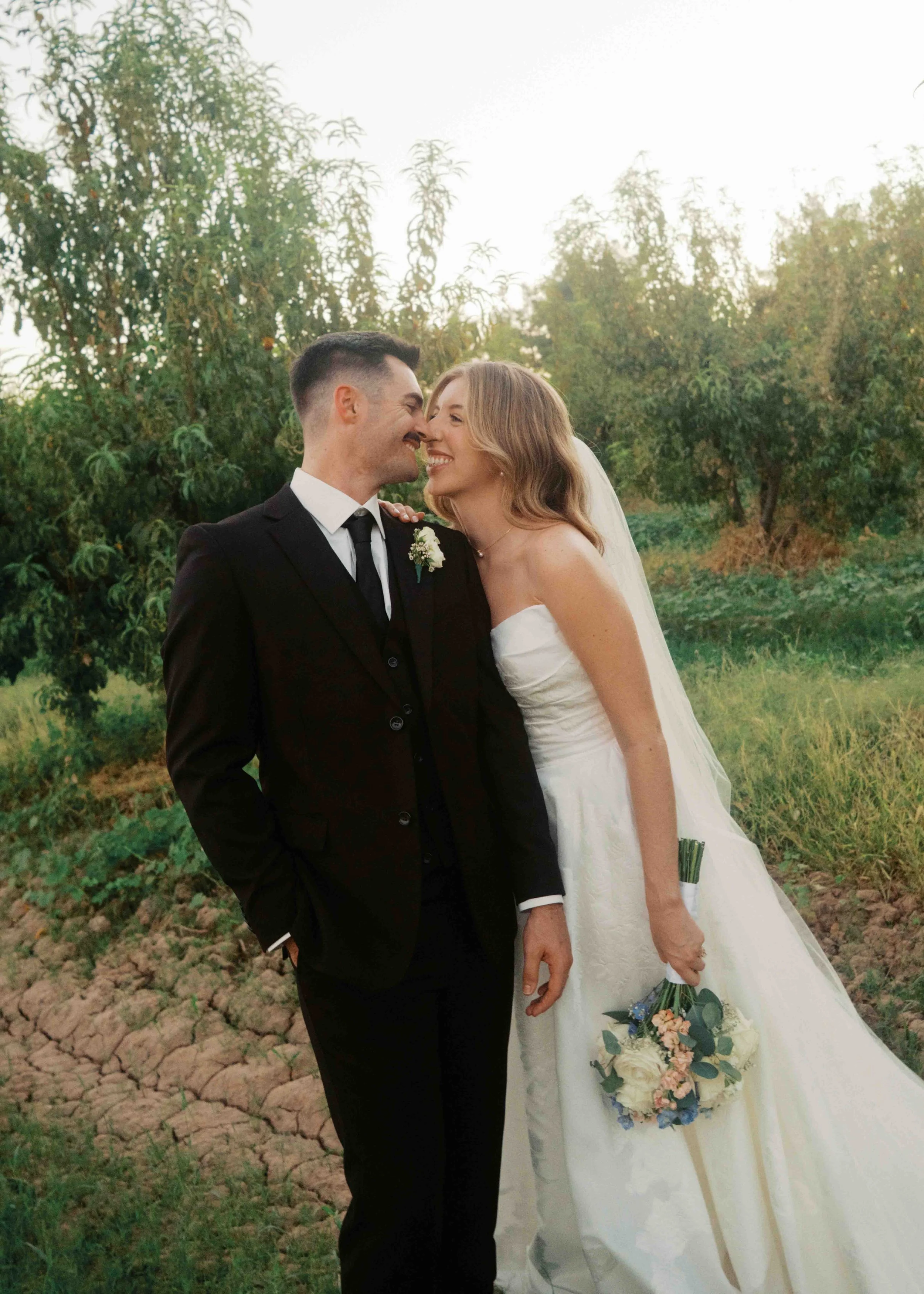 A newlywed couple smiling and leaning their noses together outdoors in a garden or field, with green trees and soil visible in the background.