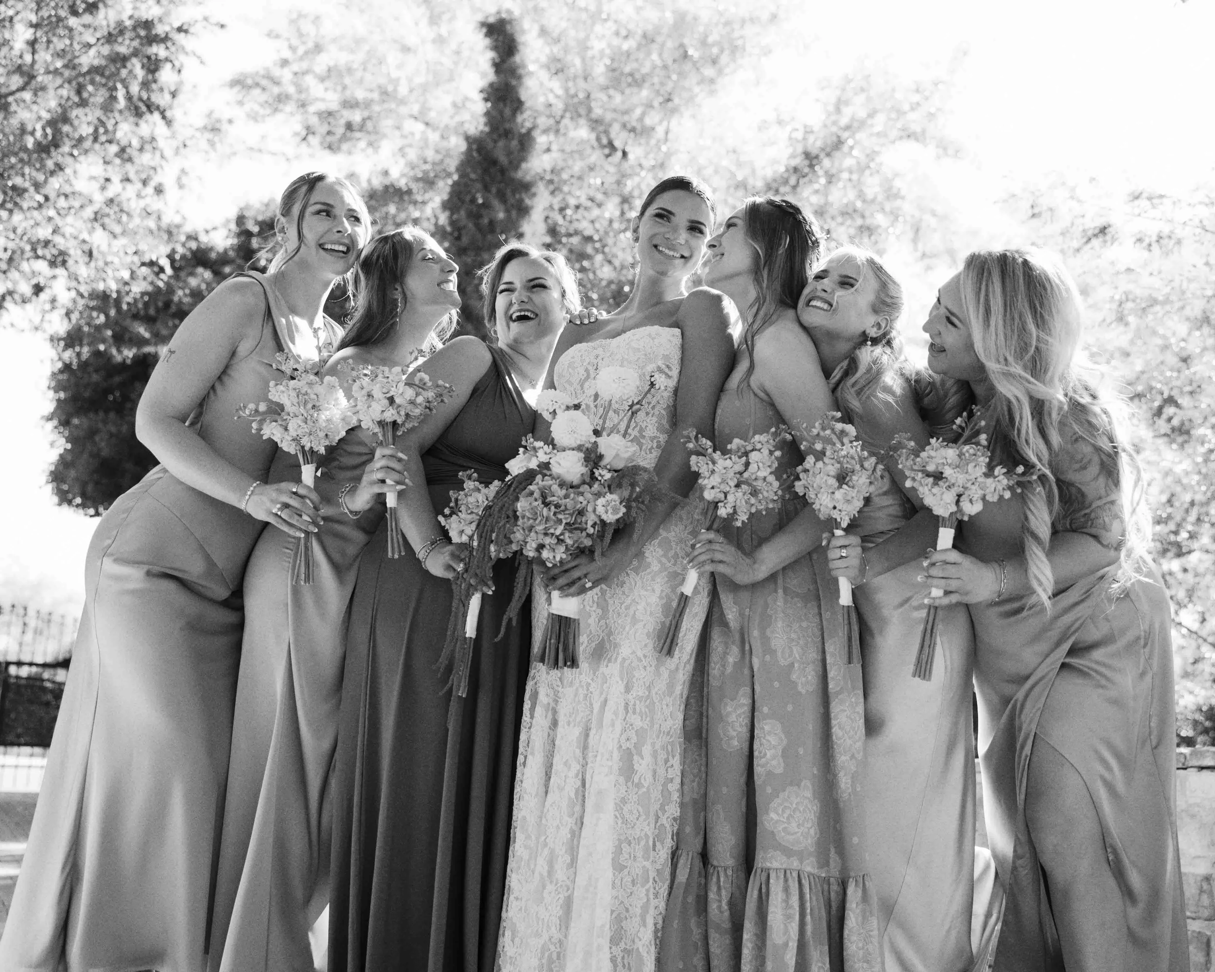 Black and white photo of a bride and six bridesmaids laughing and smiling outside during a wedding, with trees in the background.